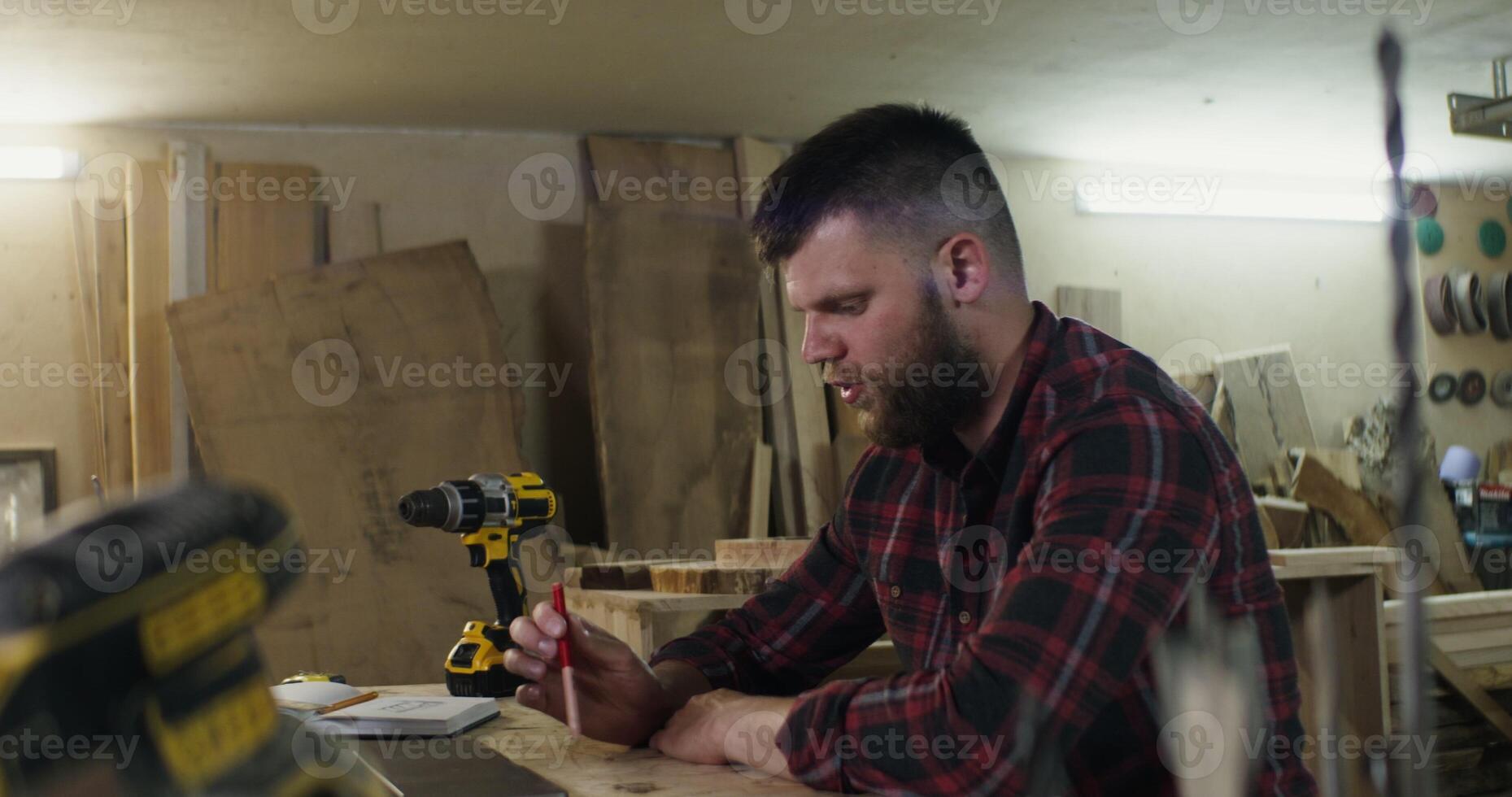 a European man with a beard and dark hair in a plaid shirt talking by call on a laptop, actively explaining something gesticulating with hands and smiles in a carpentry workshop photo