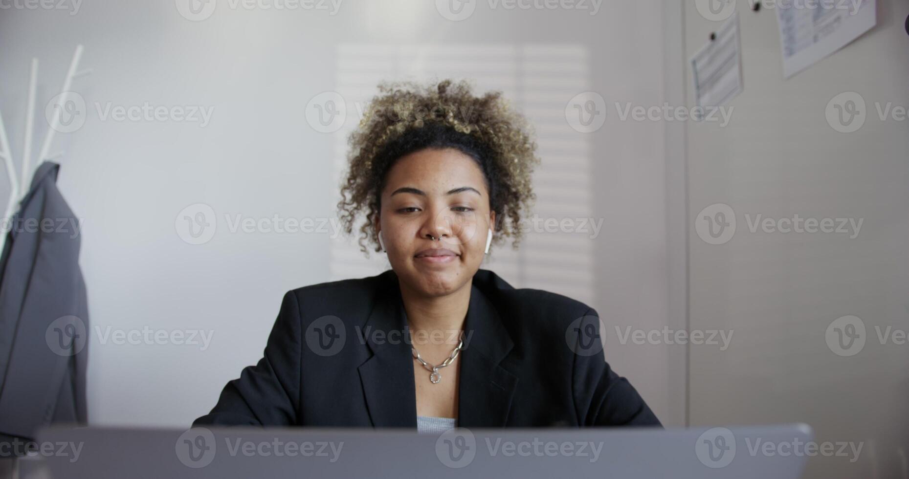 beautiful black-skinned girl with afro hairstyle in black business jacket, with earring in the nose and chain on her neck is greets and actively talking on open laptop and headphones in ears close-up photo