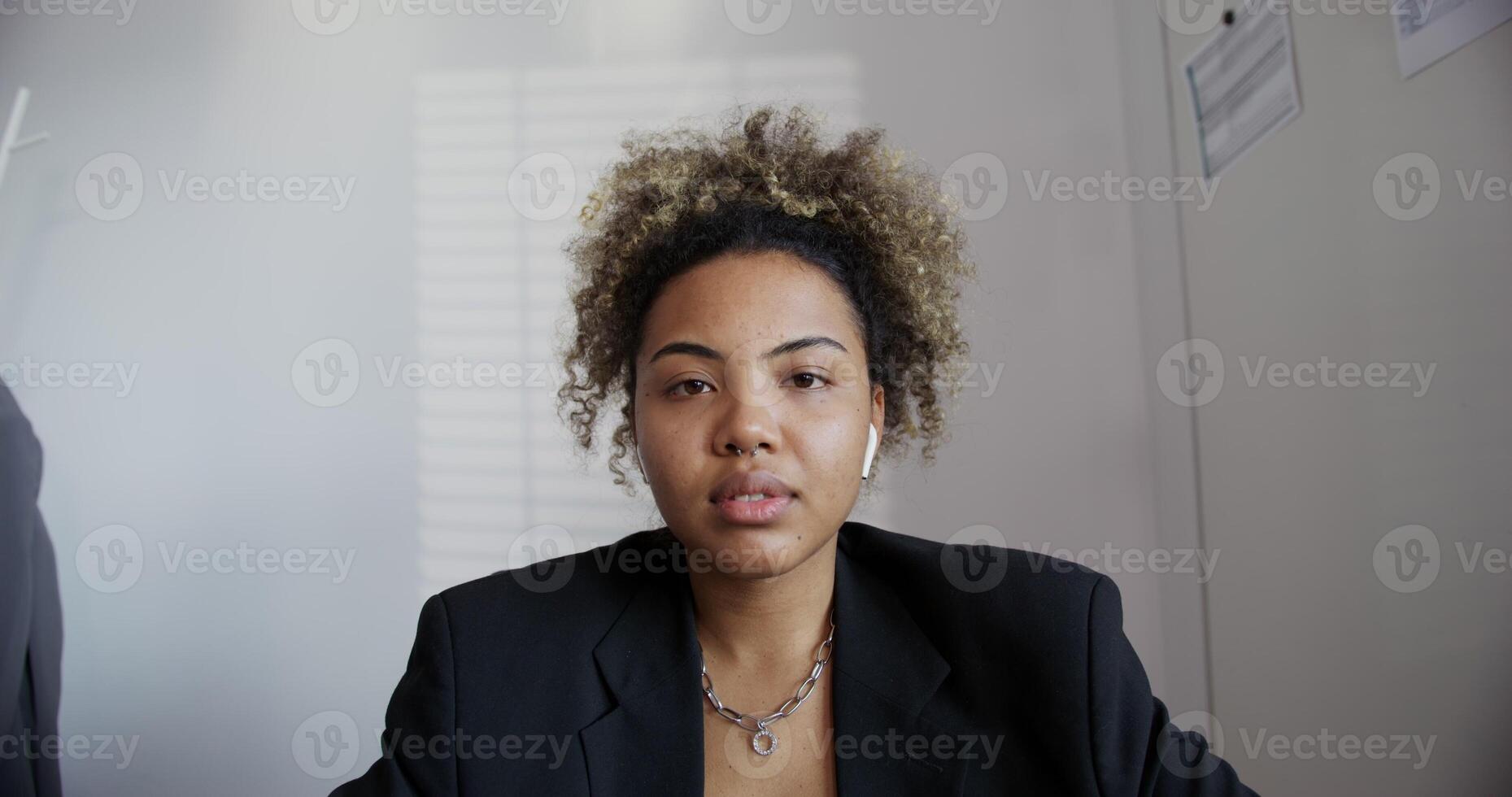 black-skinned girl with an African hairstyle in a black business jacket with, an earring in the nose and a chain around her neck with headphones in her ears is seriously looking at the camera close-up photo