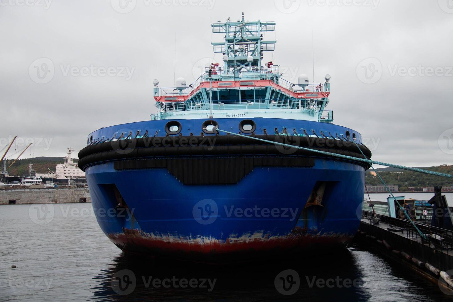 Large Blue Tugboat Docked at busy Shipyard under overcast Cloudy Sky. Visible Maritime infrastructure and Industrial background add context to Vessel's functional role in Port operations photo