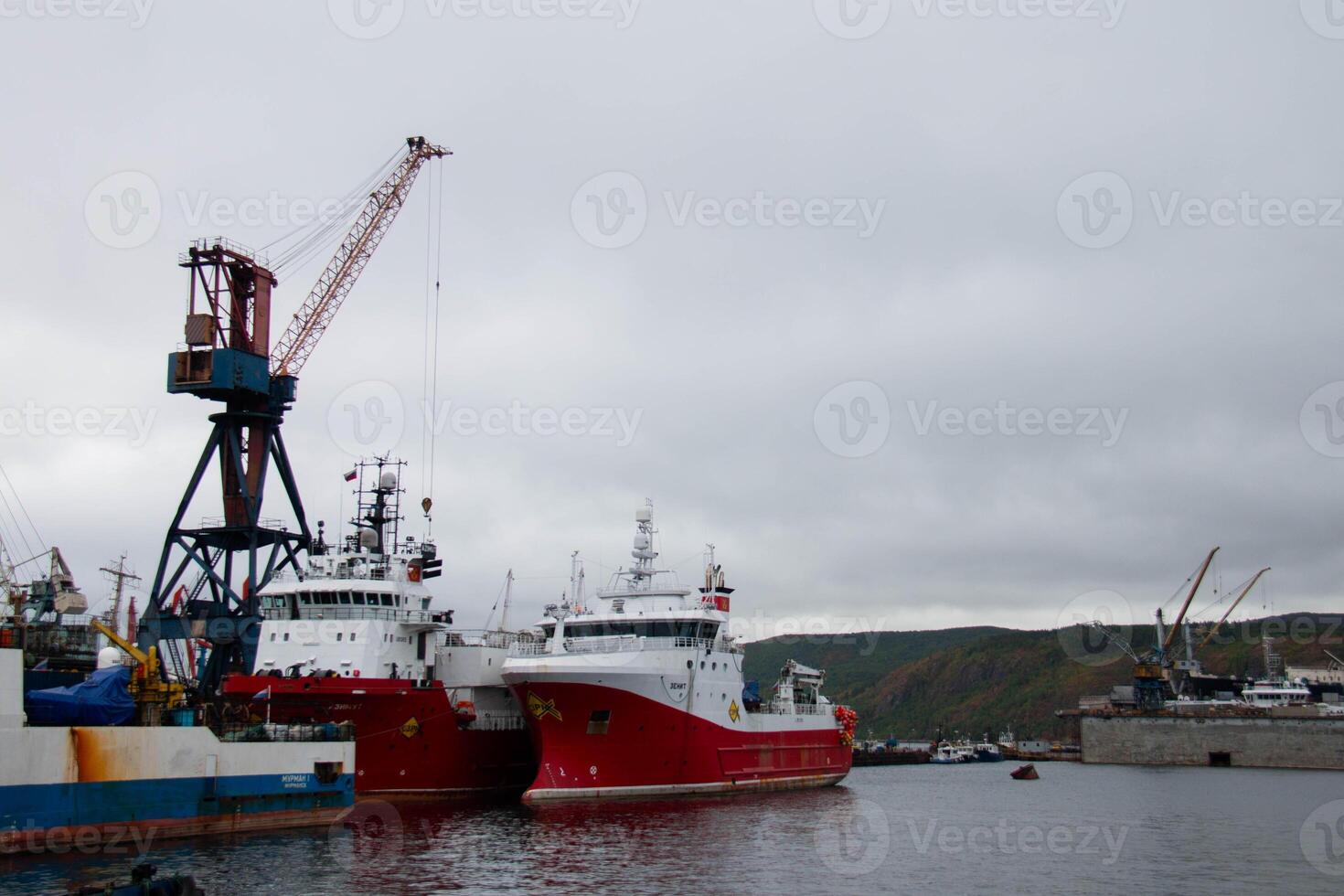 Industrial Harbor showcasing two Red Ships Docked side by side near Large crane. Overcast cloudy sky at backdrop,industrial buildings, more ships, and distant hills, a bustling maritime setting photo