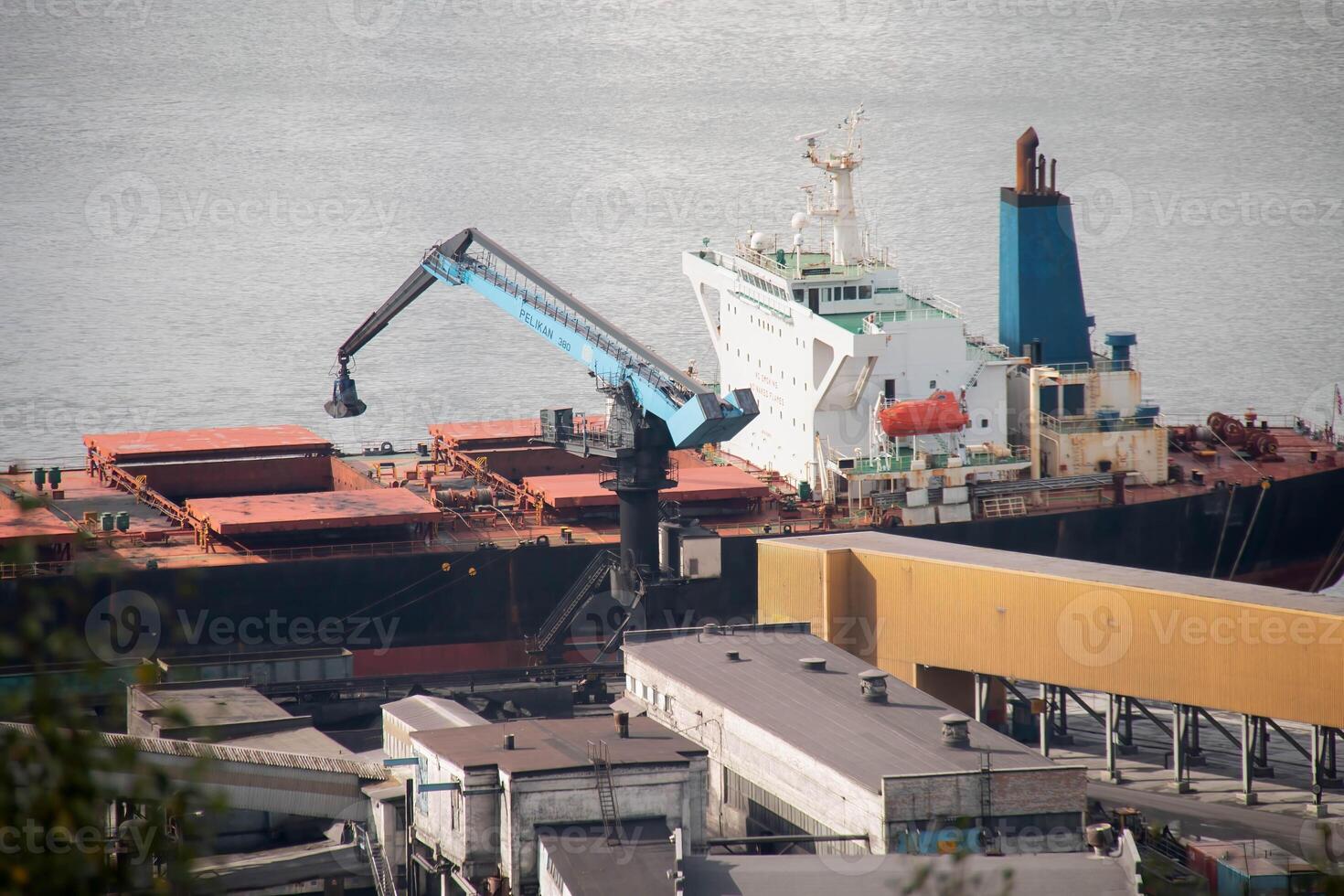Industrial Port large Cargo Ship docked Freight Terminal. Blue Gantry Crane transferring goods from Vessel. Warehouses and calm Harbor water surface background, indicating busy maritime Logistics Hub photo