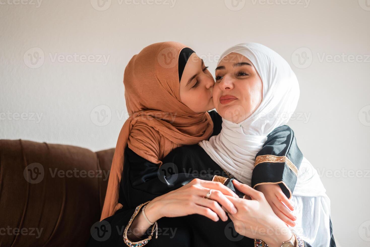Happy Muslim family at home young mother and child daughter in hijab kiss and hug with love on sofa smiling and playing together for bonding with affection on mothers day. photo