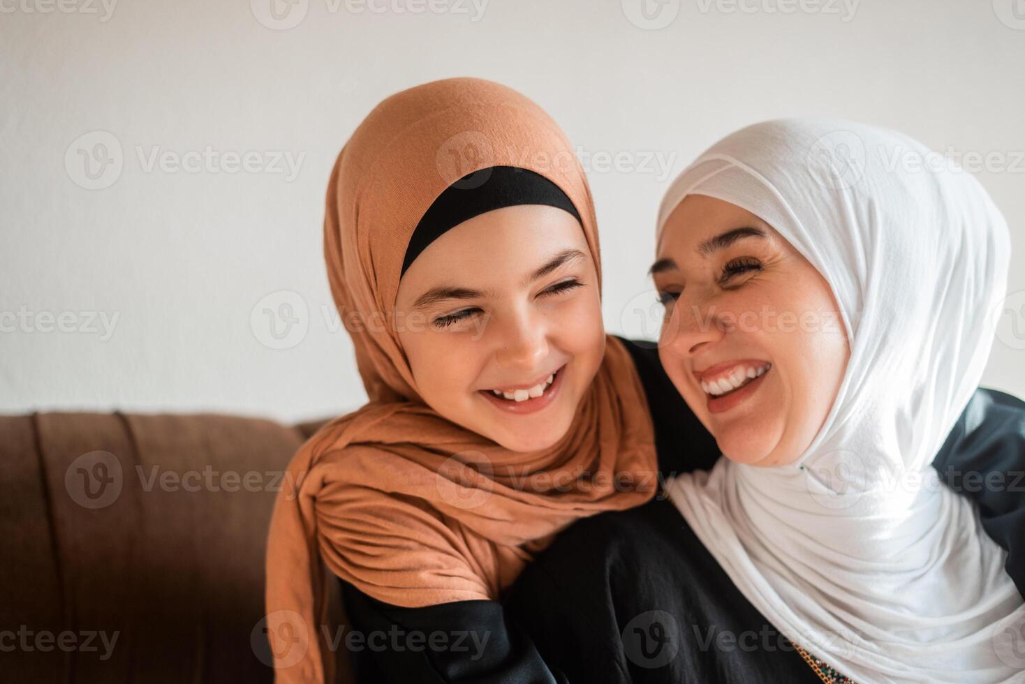 Happy Muslim family at home young mother and child daughter in hijab hug with love on sofa smiling and playing together for bonding with affection on mothers day. photo
