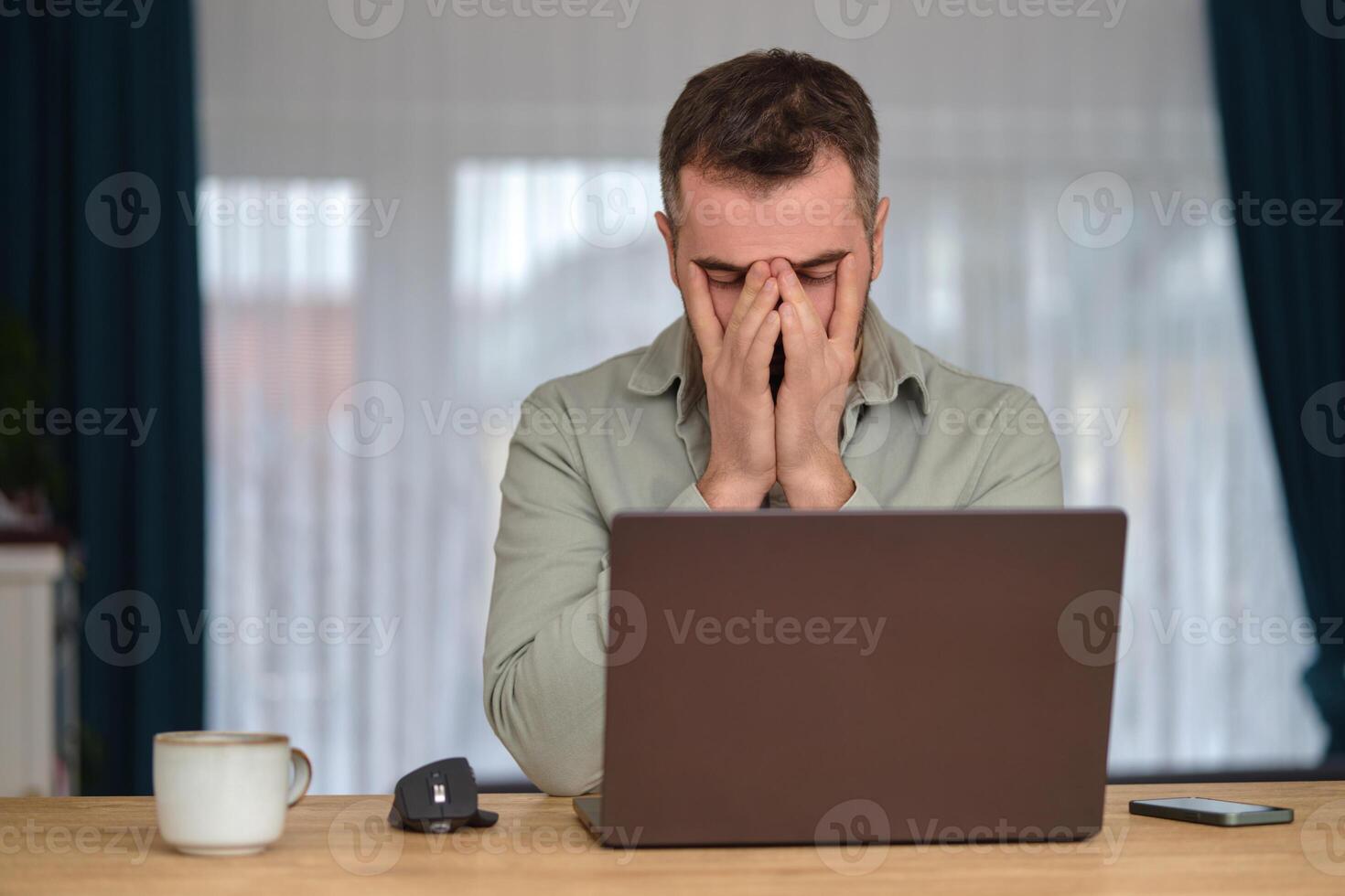 Tired man working overtime, sitting at the desk in front of a laptop and rubbing eyes with hands. photo