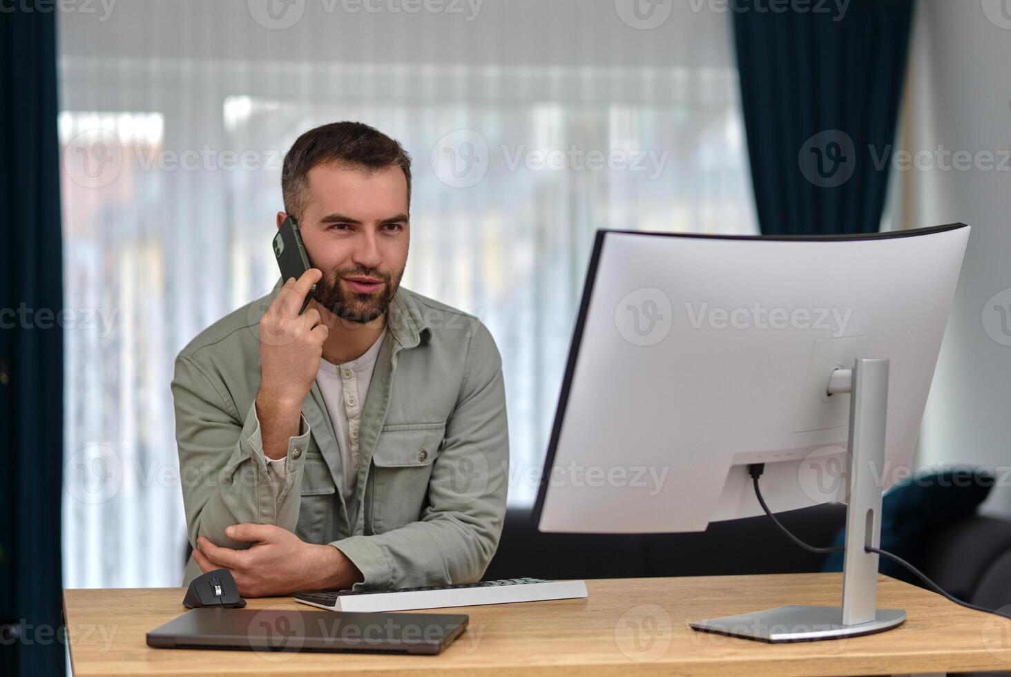 A happy man talking on his phone and using a desktop computer while working at home. photo