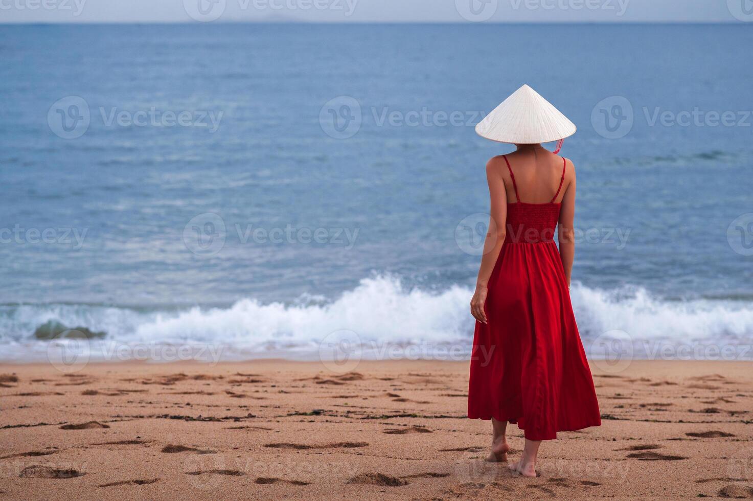 Vietnamese lady in traditional conical Non La hat and red dress on the beach against the background of the sea photo