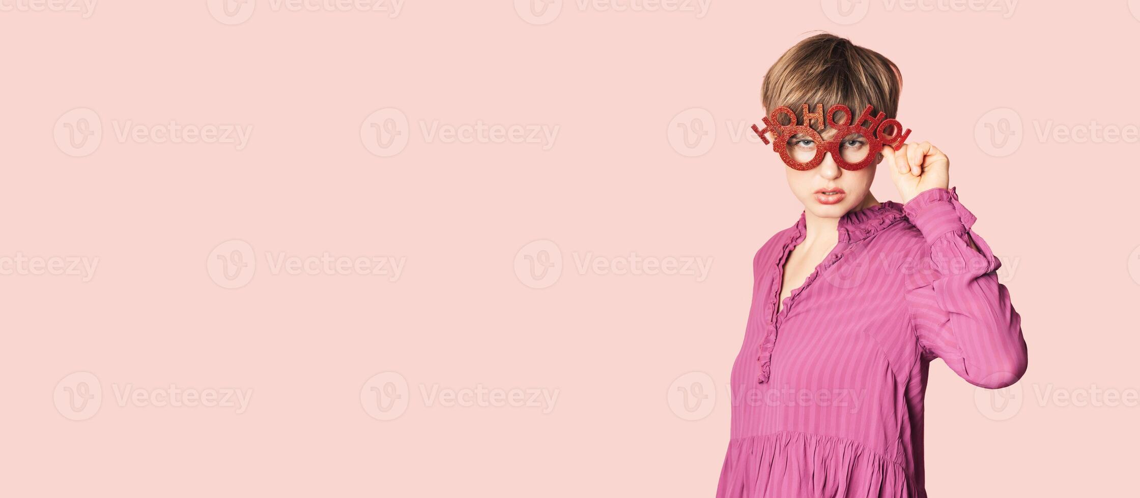 A cheerful young woman in Christmas glasses poses on a workspace layout highlighted on a simple pastel light pink background. photo