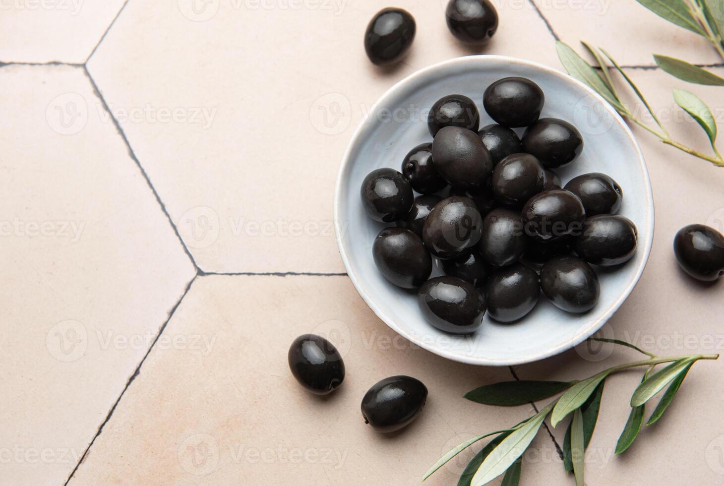 Black olives served in bowl on hexagonal beige tiles, decorated with olive tree branch photo