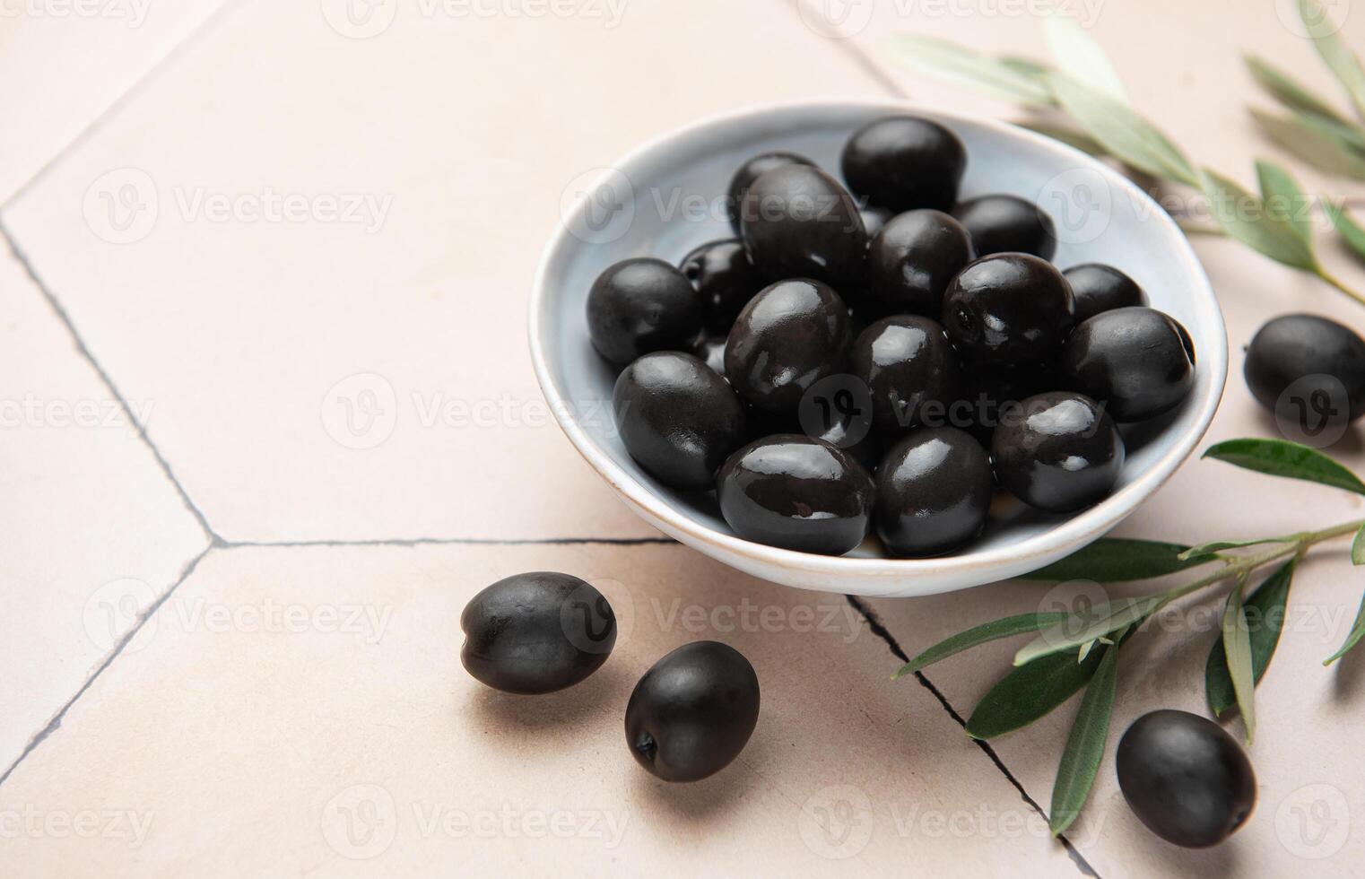 Black olives in ceramic bowl on hexagonal beige tiles, decorated with olive tree branch photo