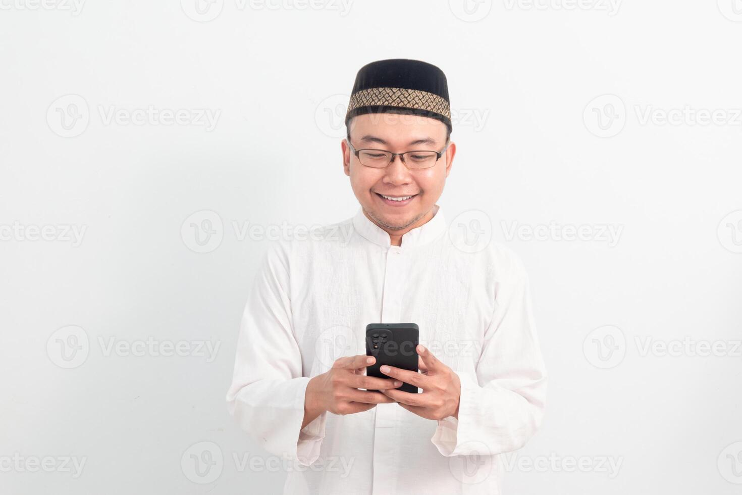 Happy Asian Muslim man wearing white clothes holding phone pose, Ramadan Eid Al Fitr and adha days celebration over white background. Islamic celebration concept. photo