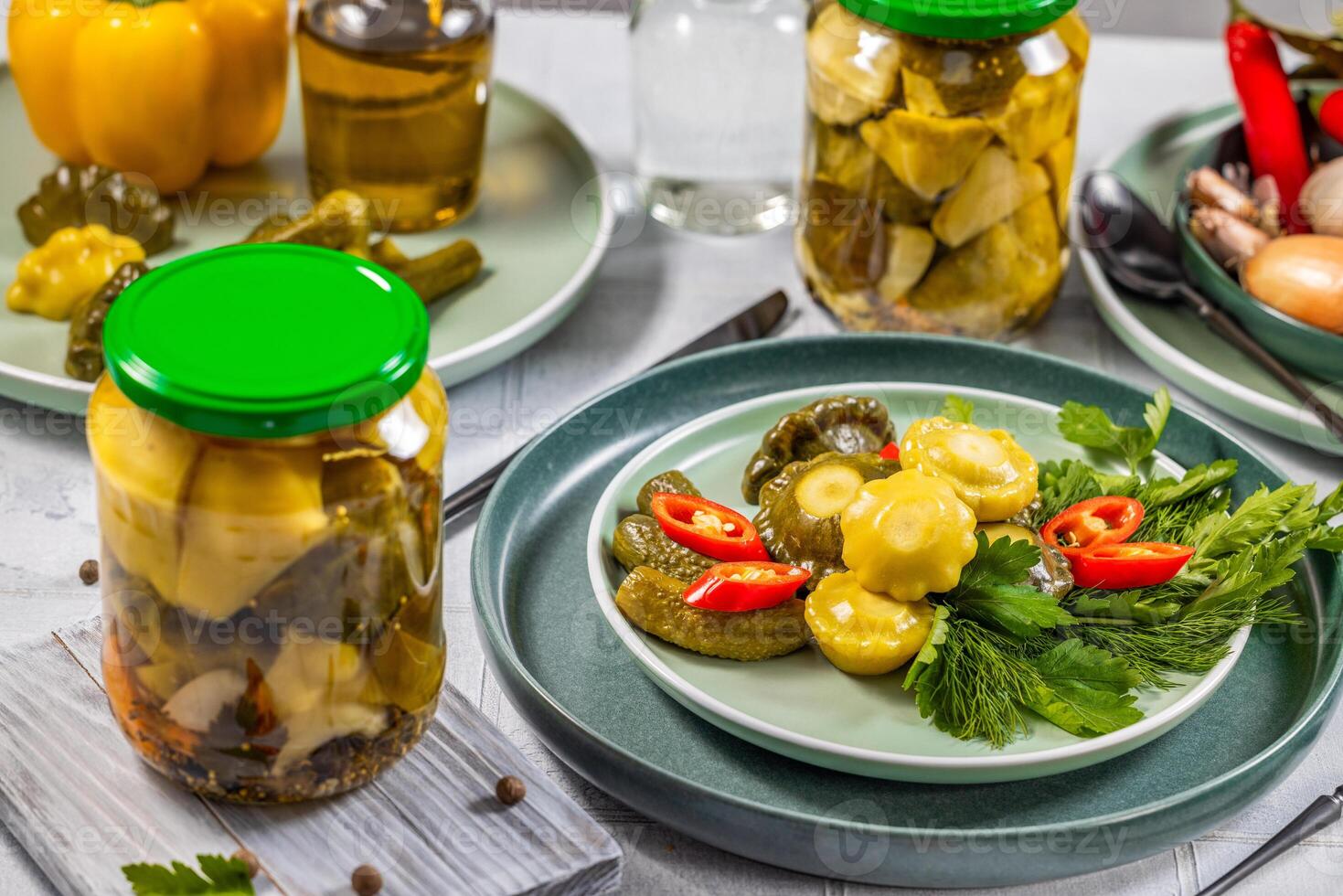 Pickled pattypan squash, gherkins, chili pepper and herbs served on a plate, with jars of pickles and other ingredients in the background photo