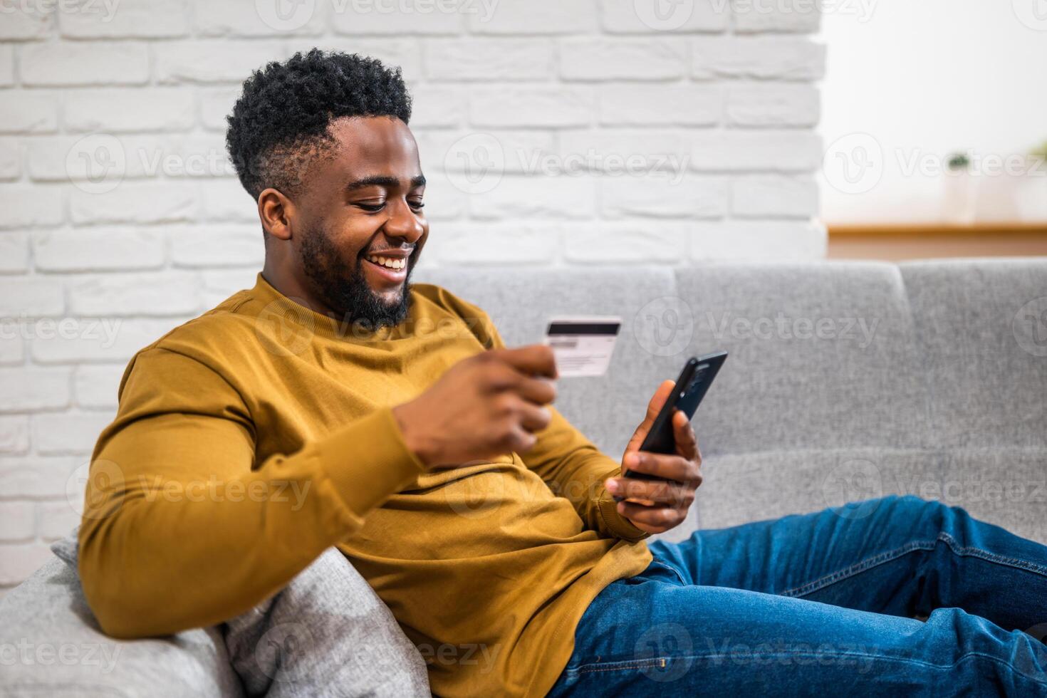 Happy young black man using credit card and mobile phone while enjoying sitting on sofa at his cozy home. photo