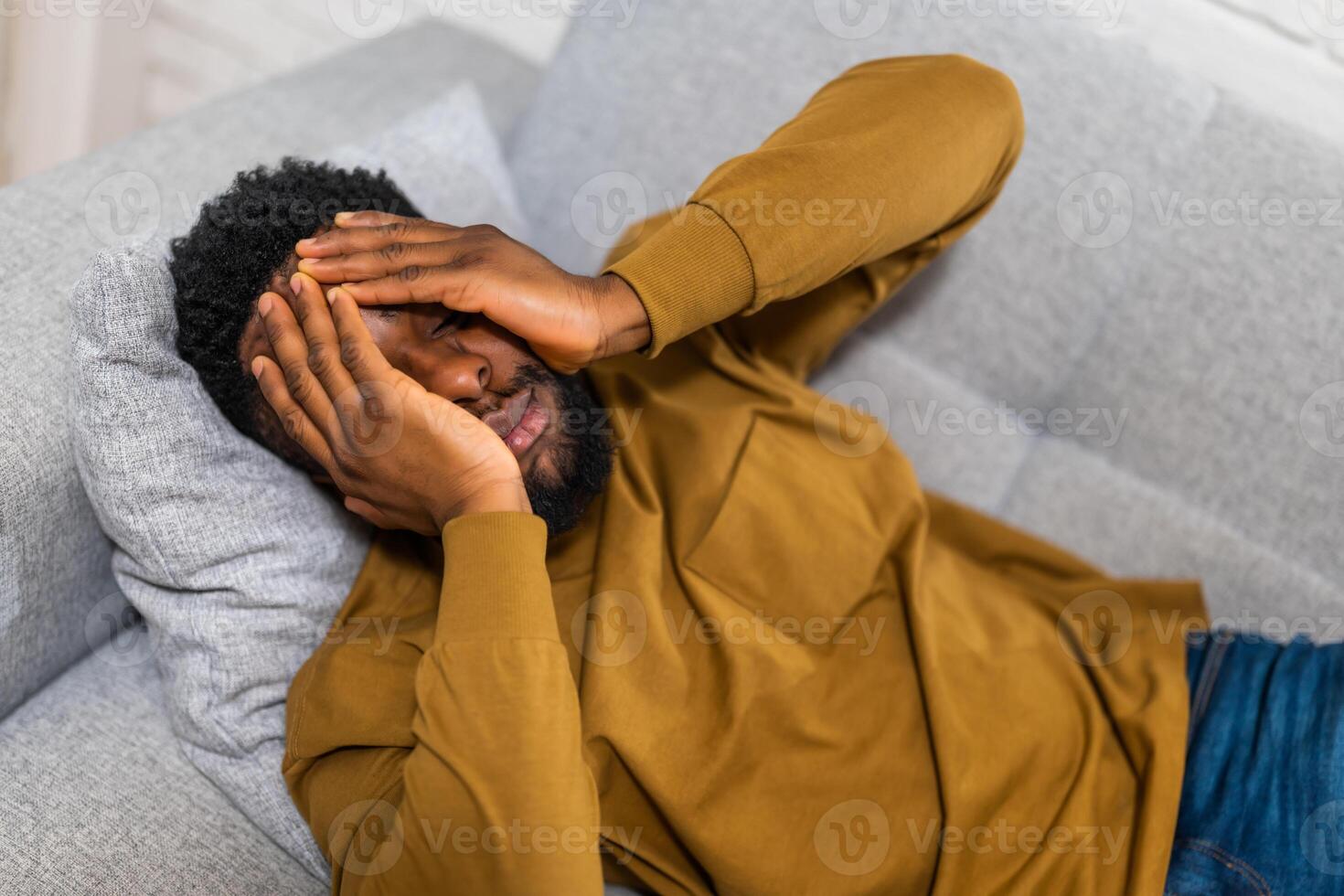 Exhausted and depressed African American man holding head in hands and lying on sofa at his home. photo