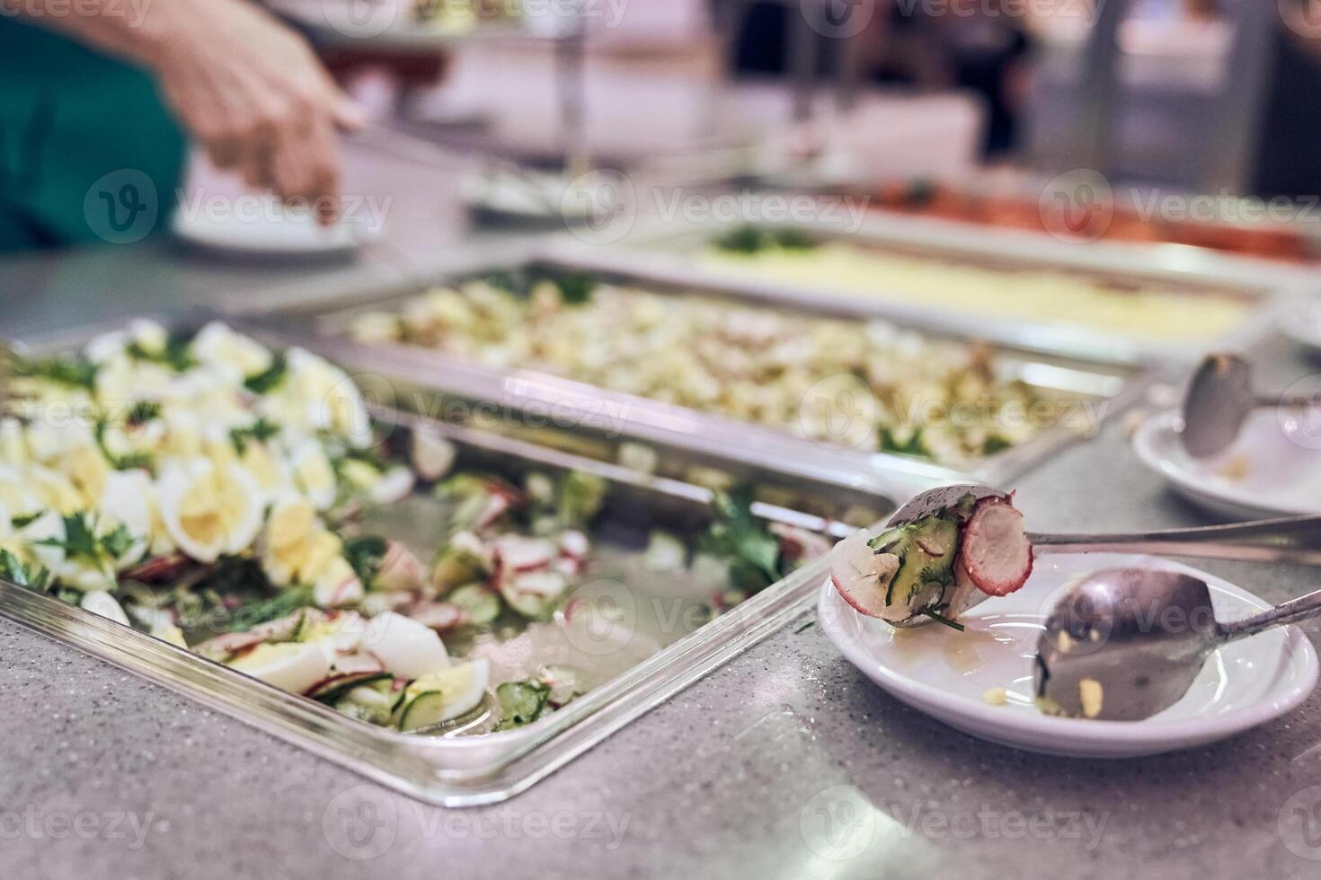 Selective focus on the hand of an unrecognizable volunteer who is about to put nutritious food on a plate in a modern charity canteen photo