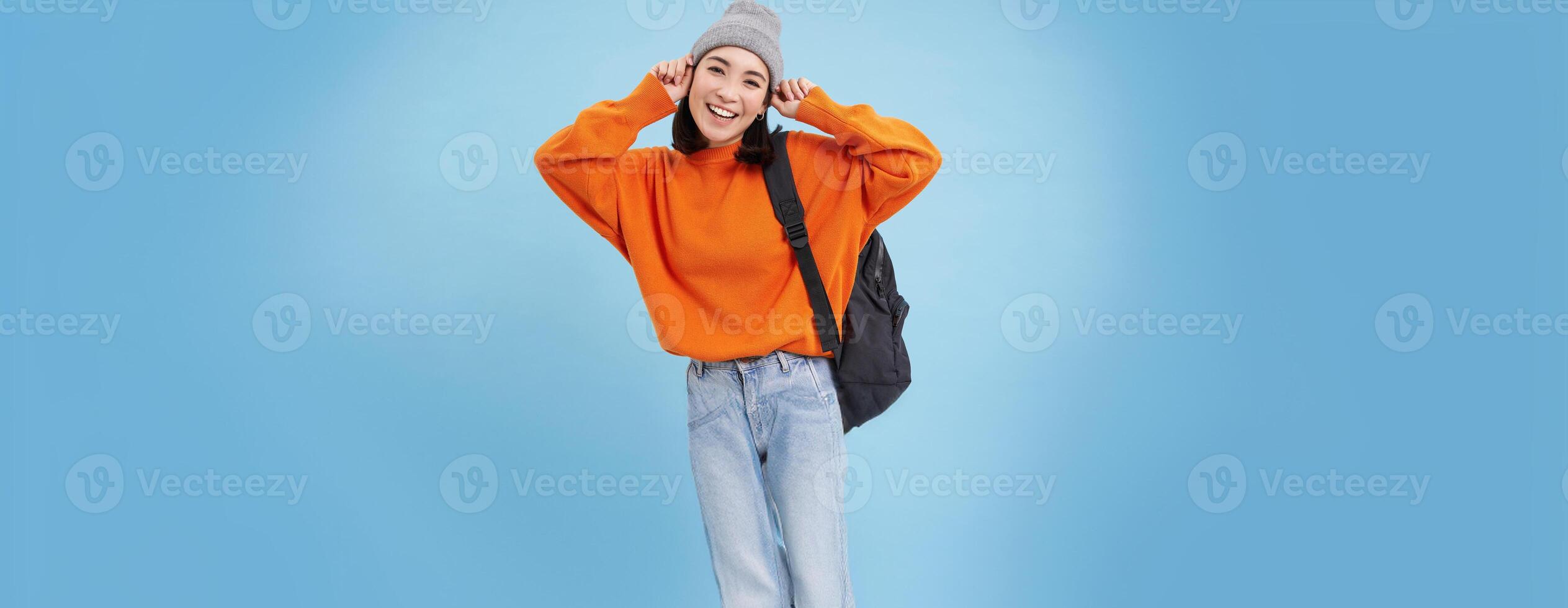Stylish young asian woman in warm hat, walking with backpack, going somewhere with bag, standing over blue studio background photo