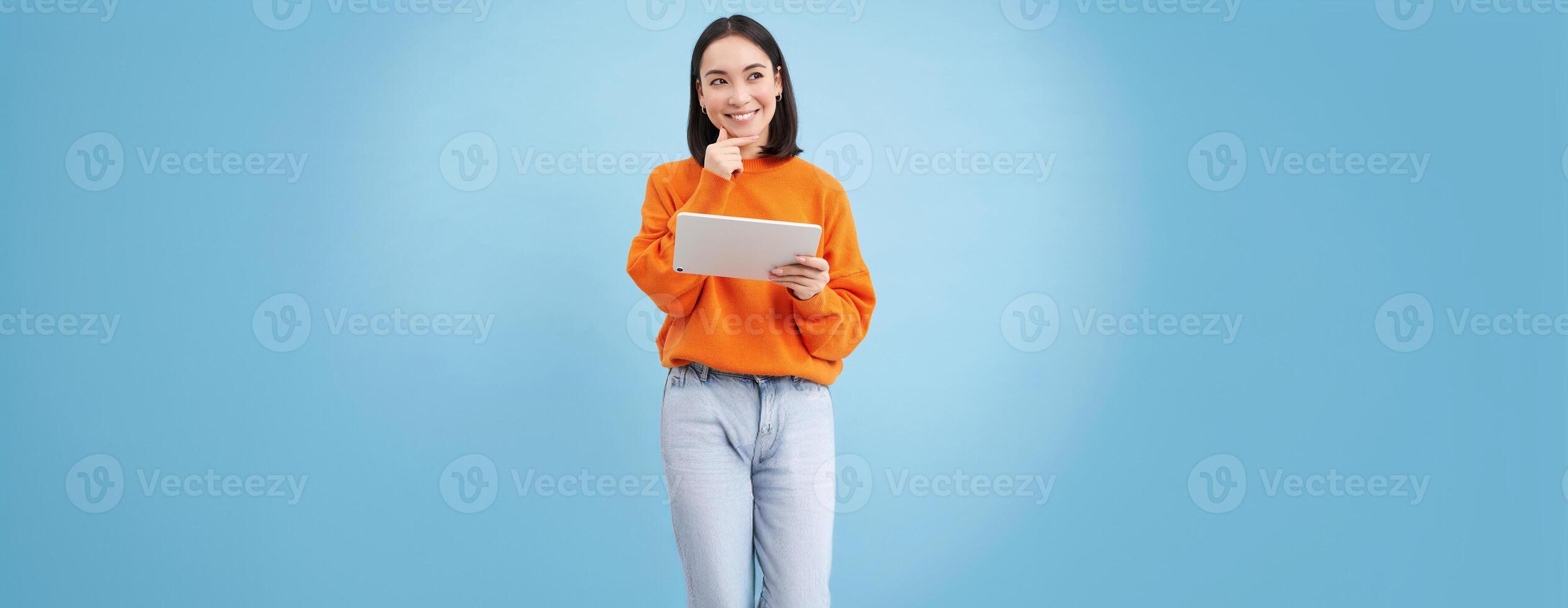 Woman with digital tablet thinking, looking aside with thoughtful face expression, standing over blue background photo