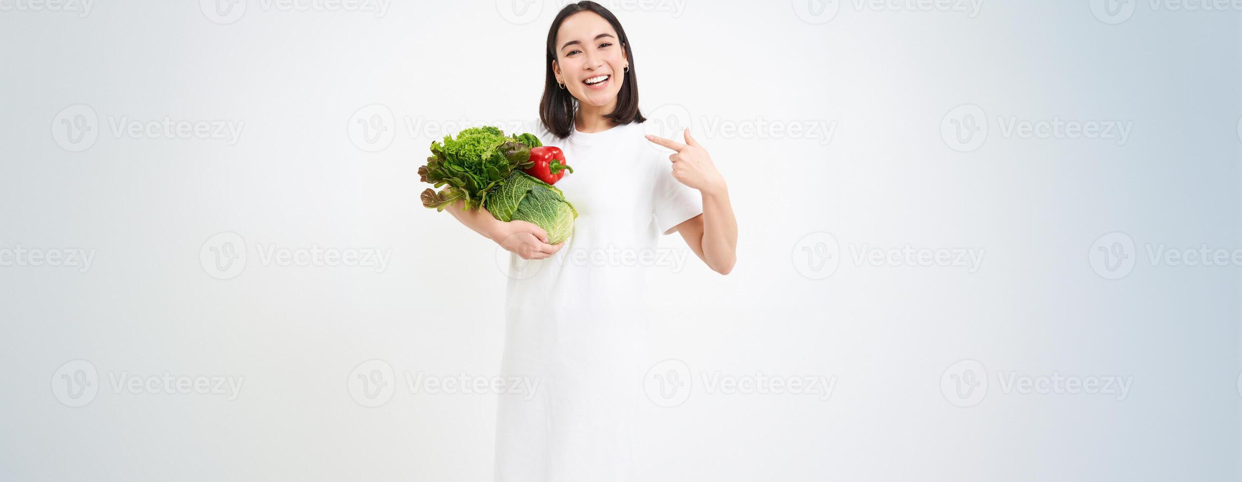 Image of happy korean woman, pointing at vegetables, smiling, recommending eating healthy raw food, green diet concept, white background photo