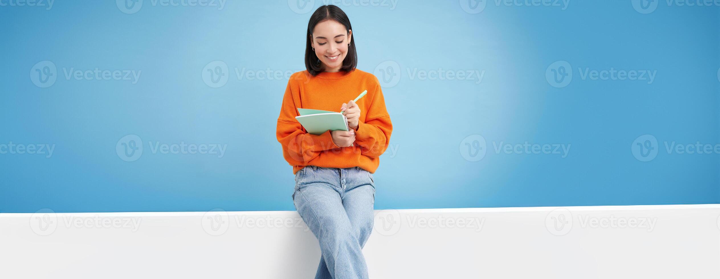 Smiling cute girl student, making notes, writing down homework, studying, standing over blue studio background photo