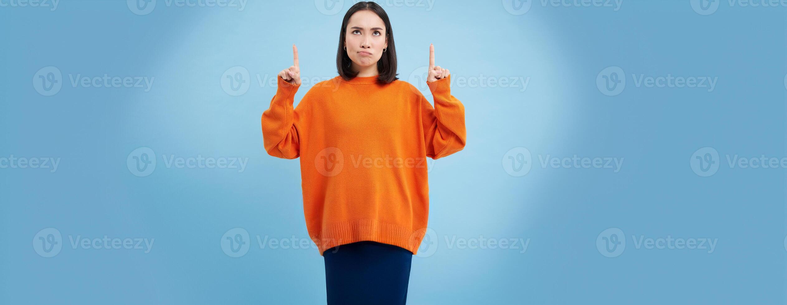 Perplexed asian girl, looks and points up with frustrated, thinking face expression, shows advertisement and reads it with doubt, stands over blue background photo