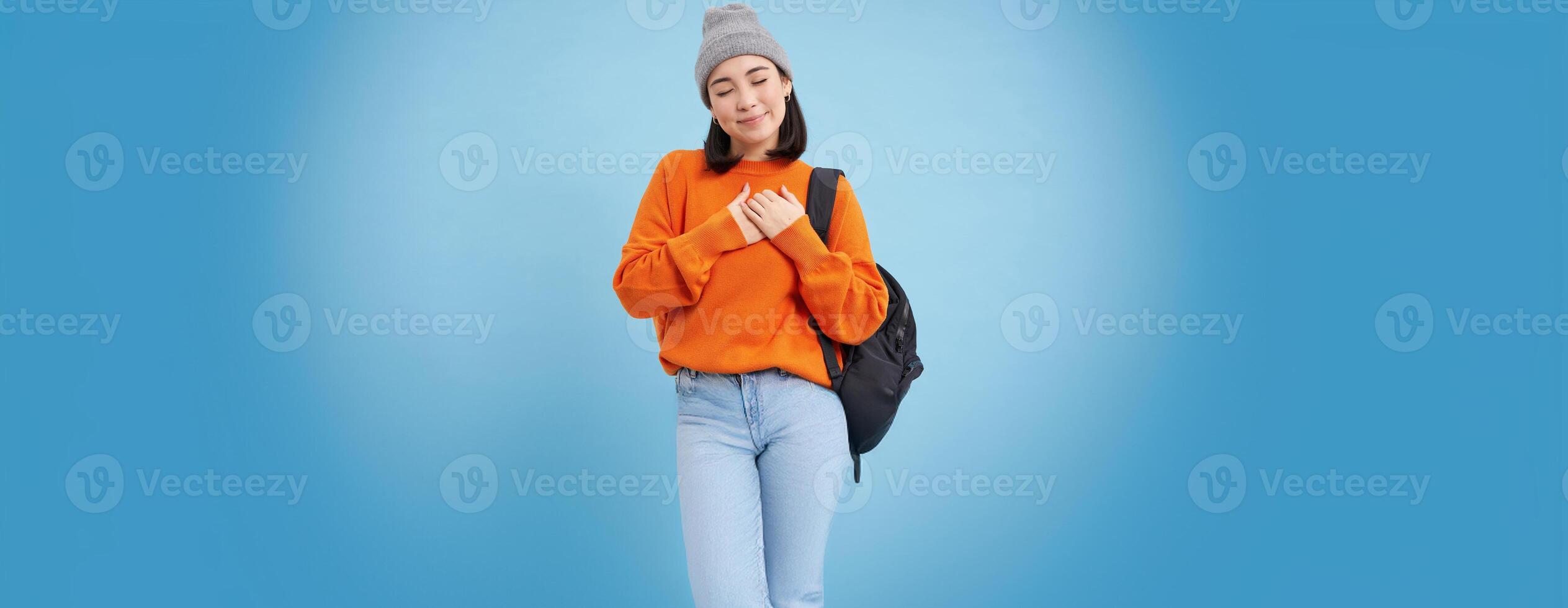 Romantic young woman in hat, holds backpack on shoulder, holds hands on heart and closes eyes, dreaming of something, standing over blue background photo