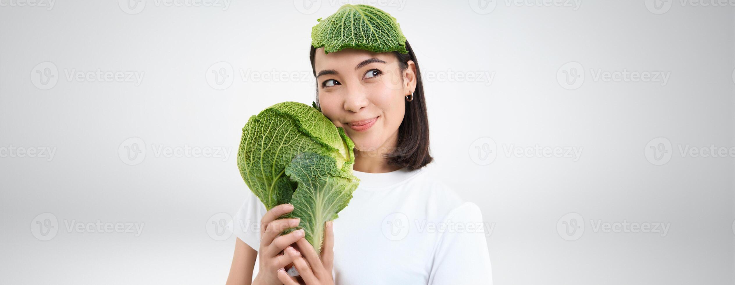 Close up portrait of beautiful and healthy vegan woman, holding lettuce, has cabbage lead on head, white background photo