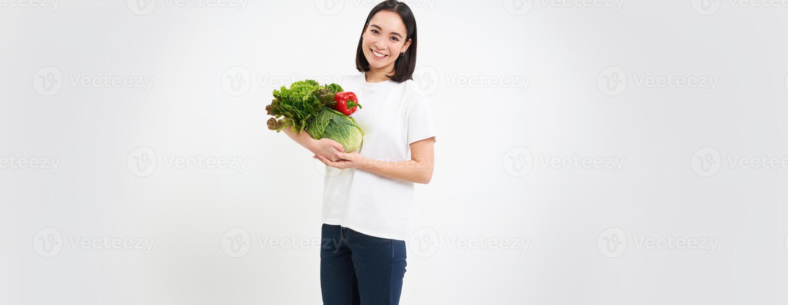 Portrait of beautiful smiling korean woman, holds bouquette of vegetables, buying green organic food at market, white background photo