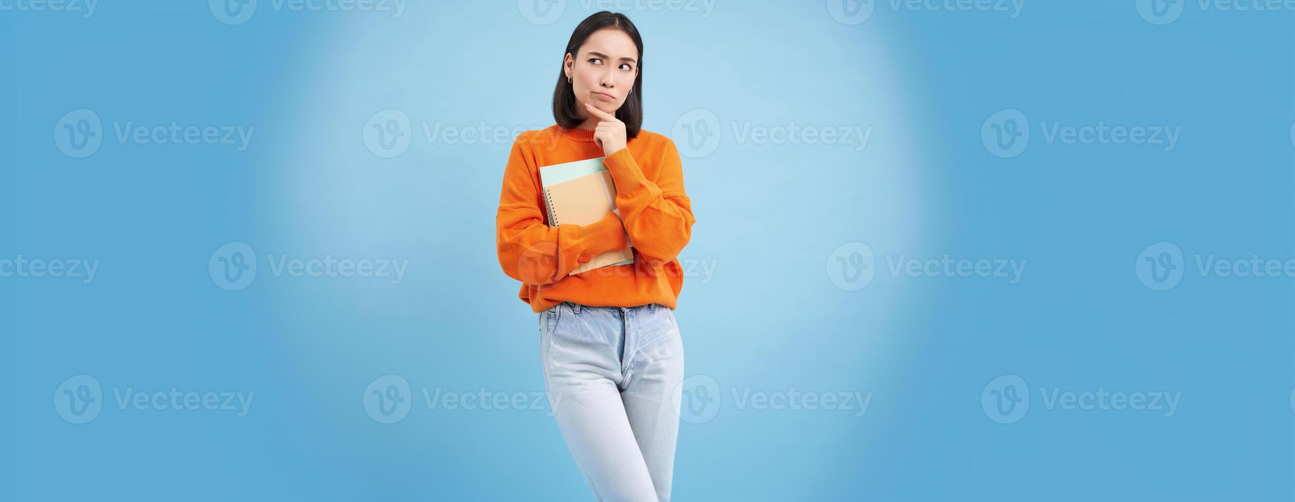 Asian woman standing with notebooks, thinking, looking aside with thoughtful face expression, standing over blue background photo