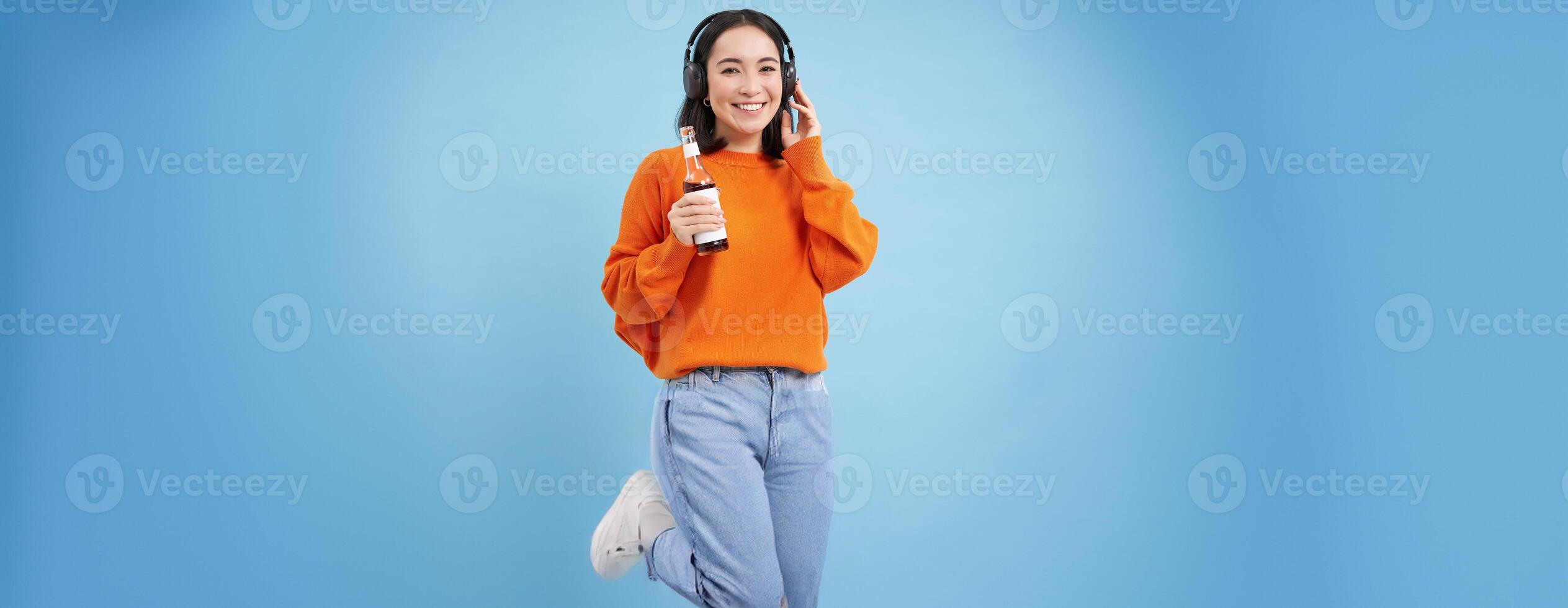 Beautiful asian woman listens music in headphones, drinks coke from bottle and smiles at camera, blue background photo