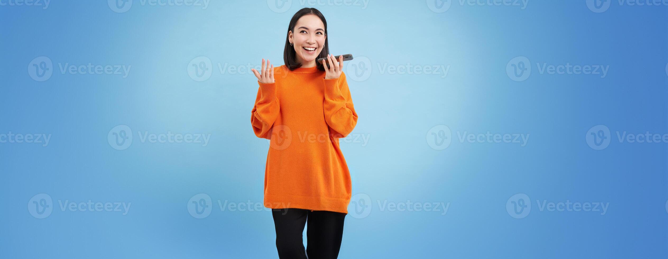 Cheerful smiling korean woman talk on speakerphone, records voice message, learns how to speak using language learning mobile app, blue background photo