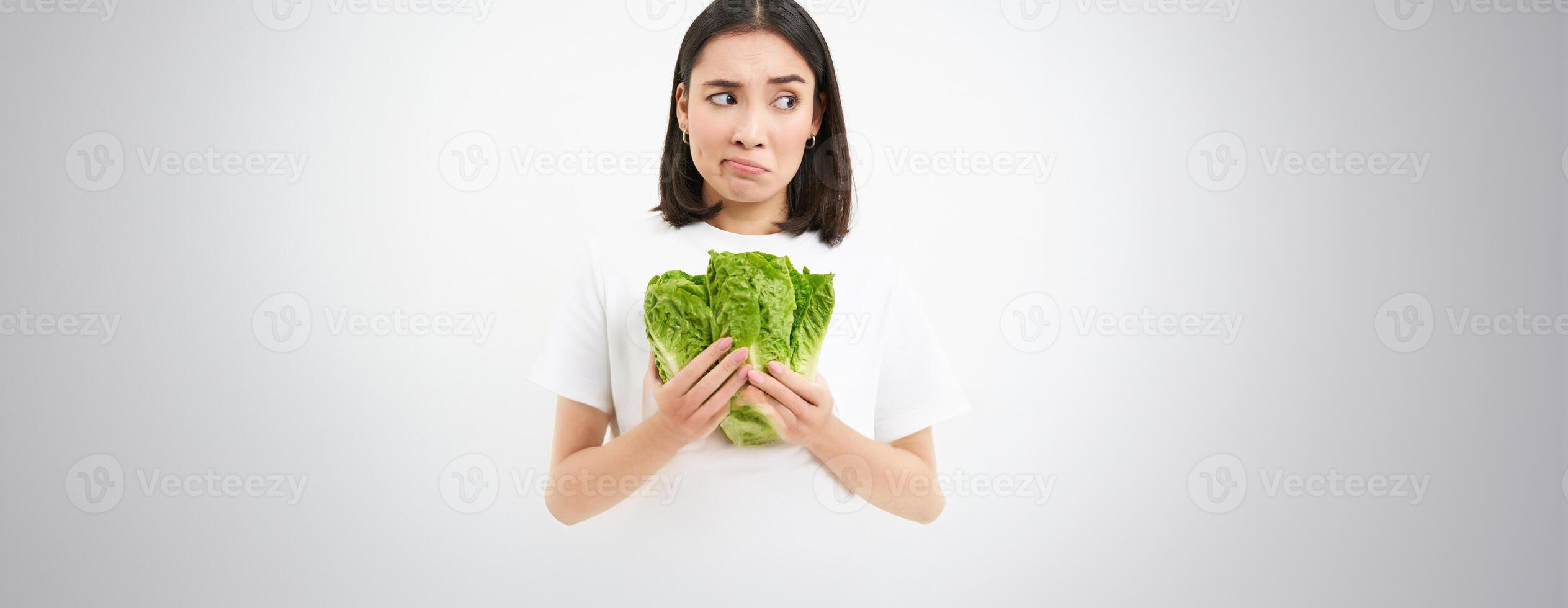 Healthy diet and raw food. Sad and unhappy asian woman, holding fresh lettuce leaves, cabbage in hands, eating vegetables, white background photo
