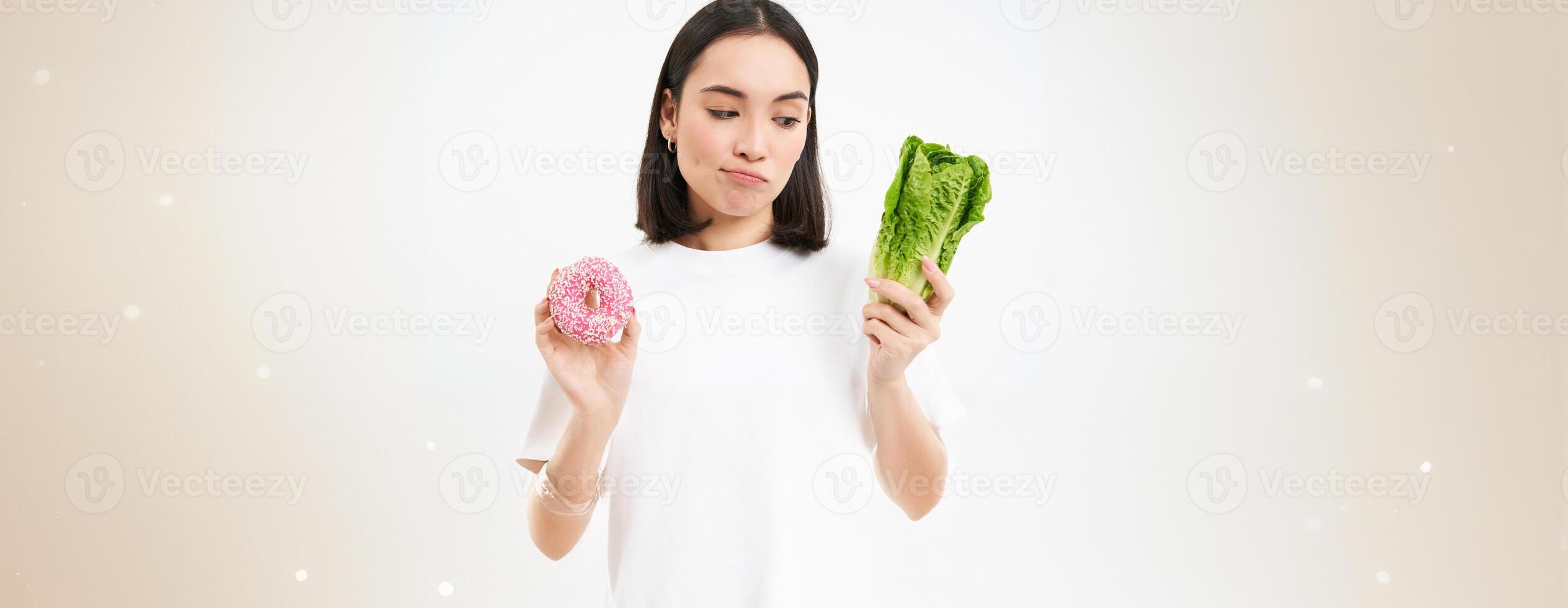 Healthy lifestyle concept. Young woman deciding between healthy vegetables, cabbage and delicious glazed doughnuts, white background photo