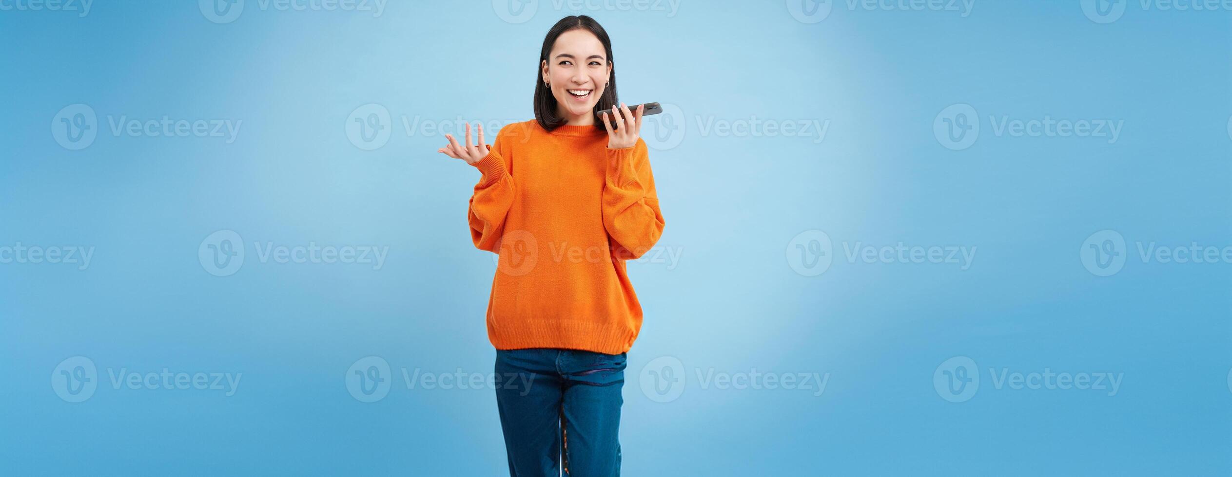 Happy asian woman records voice message, repeats words in language learning mobile app, talks in smartphone speakerphone, blue background photo