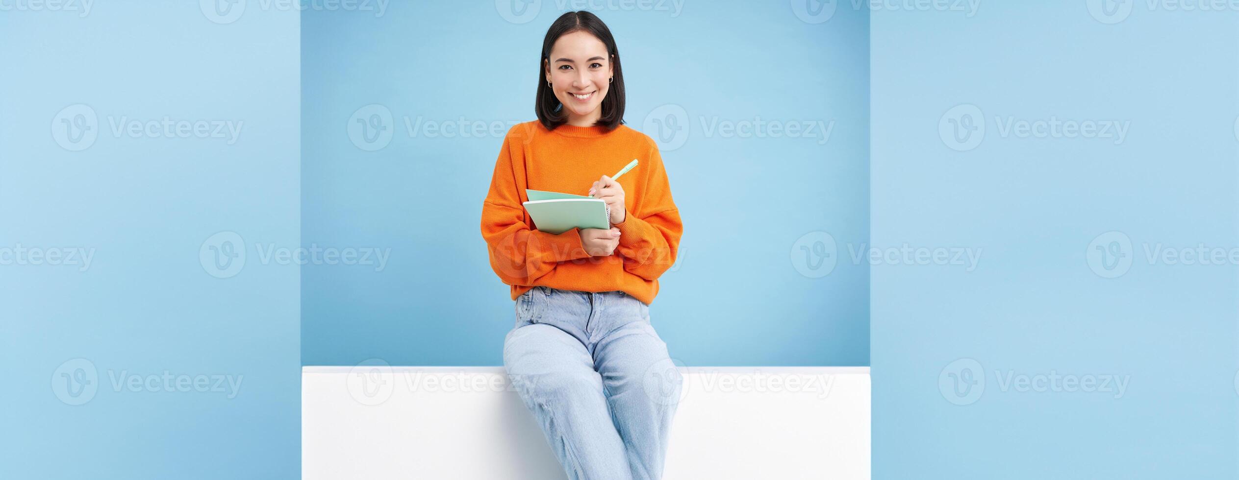 Smiling cute girl student, making notes, writing down homework, studying, standing over blue studio background photo