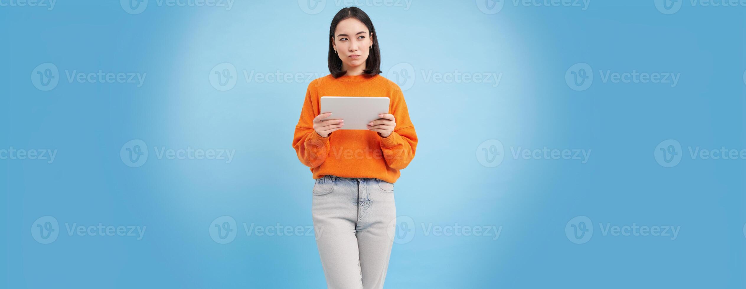 Woman with digital tablet thinking, looking aside with thoughtful face expression, standing over blue background photo