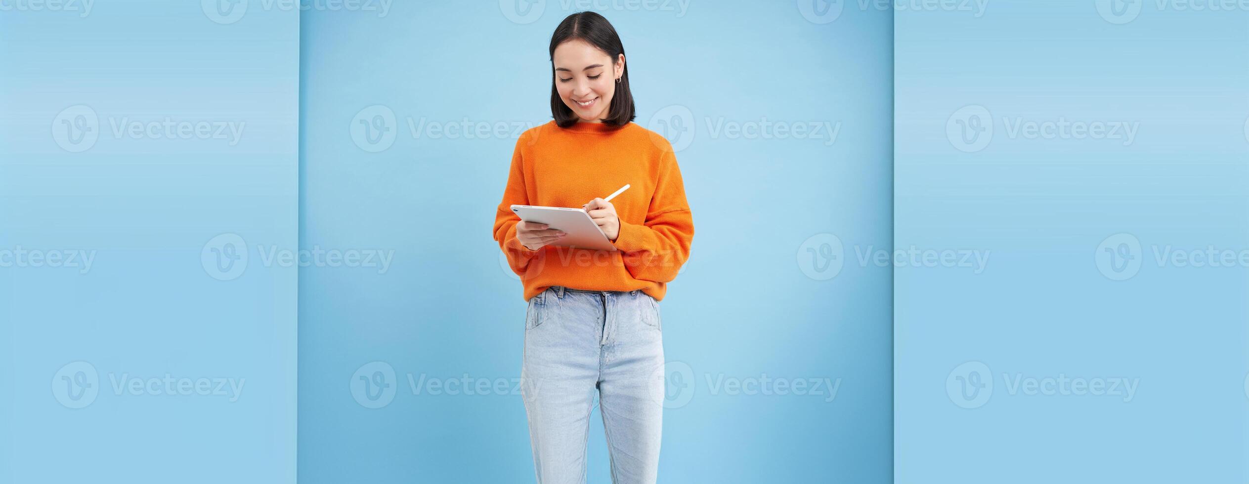 Portrait of smiling female tutor, girl student making notes on digital tablet, drawing in app, standing over blue background photo