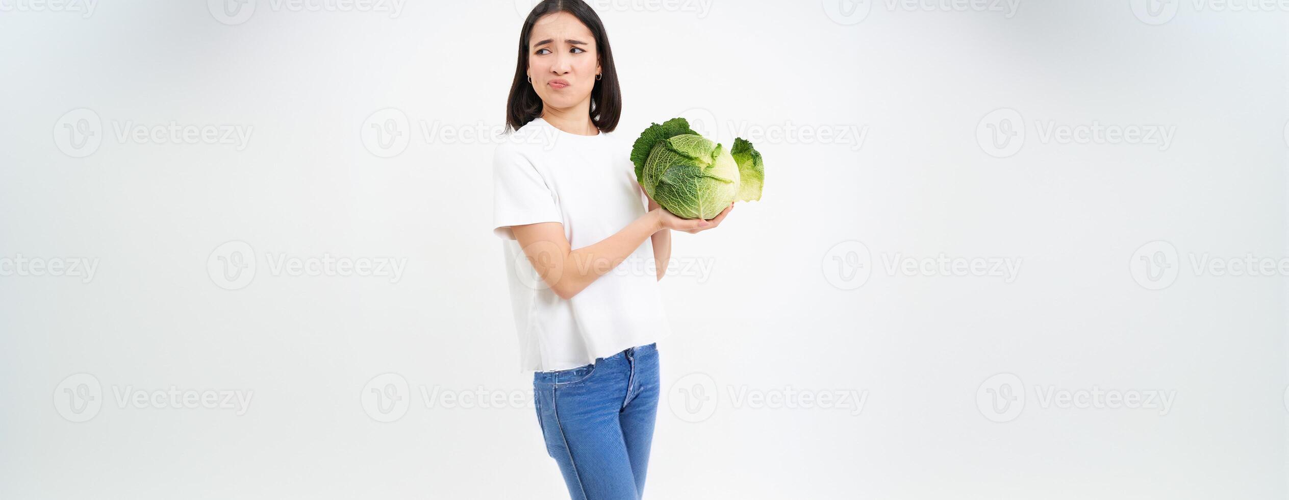 Asian woman looks with disgust and dislike at cabbage, hates vegetables, isolated on white background photo