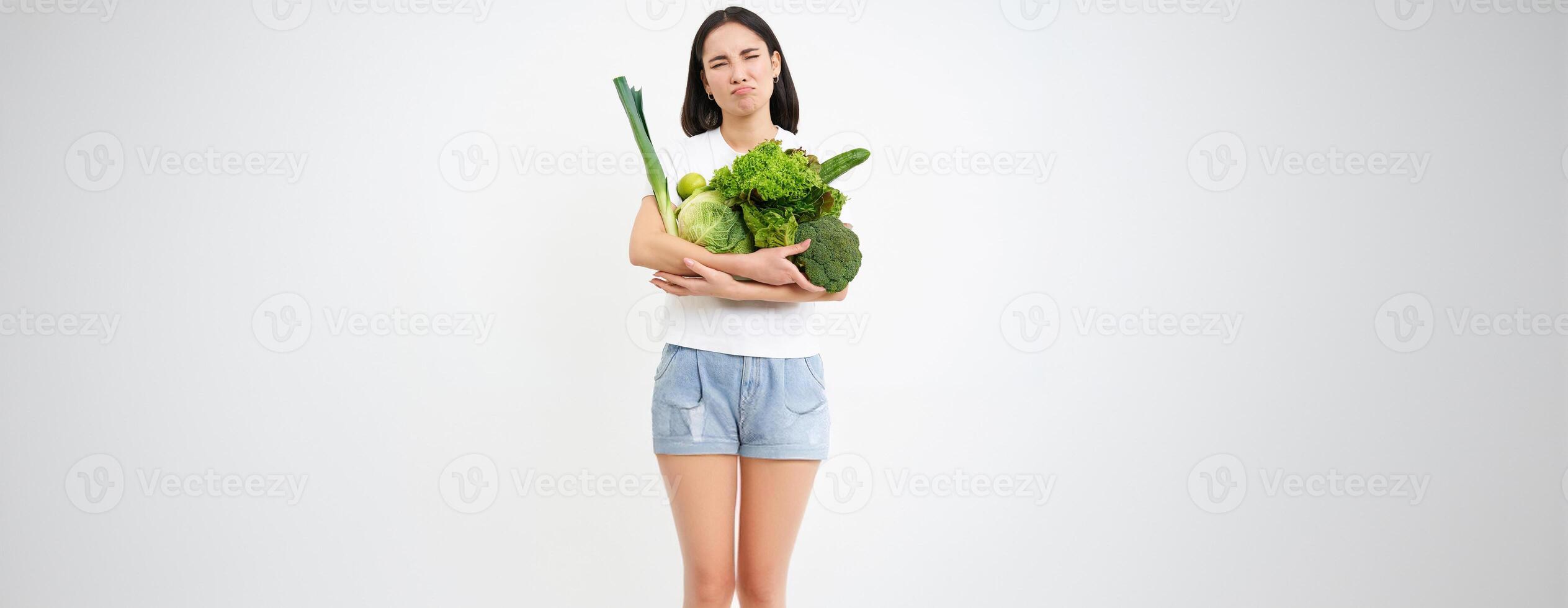 Woman with disgusted face, holding vegetables, green organic food, hates diets and detox, white background photo