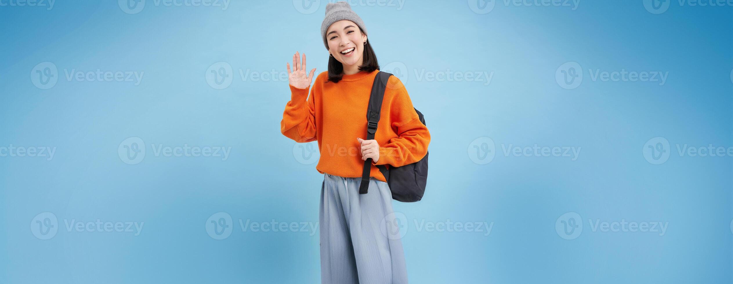 Friendly girl says hello, holds backpack and waves hand, greets you and laughs, stands over blue background photo