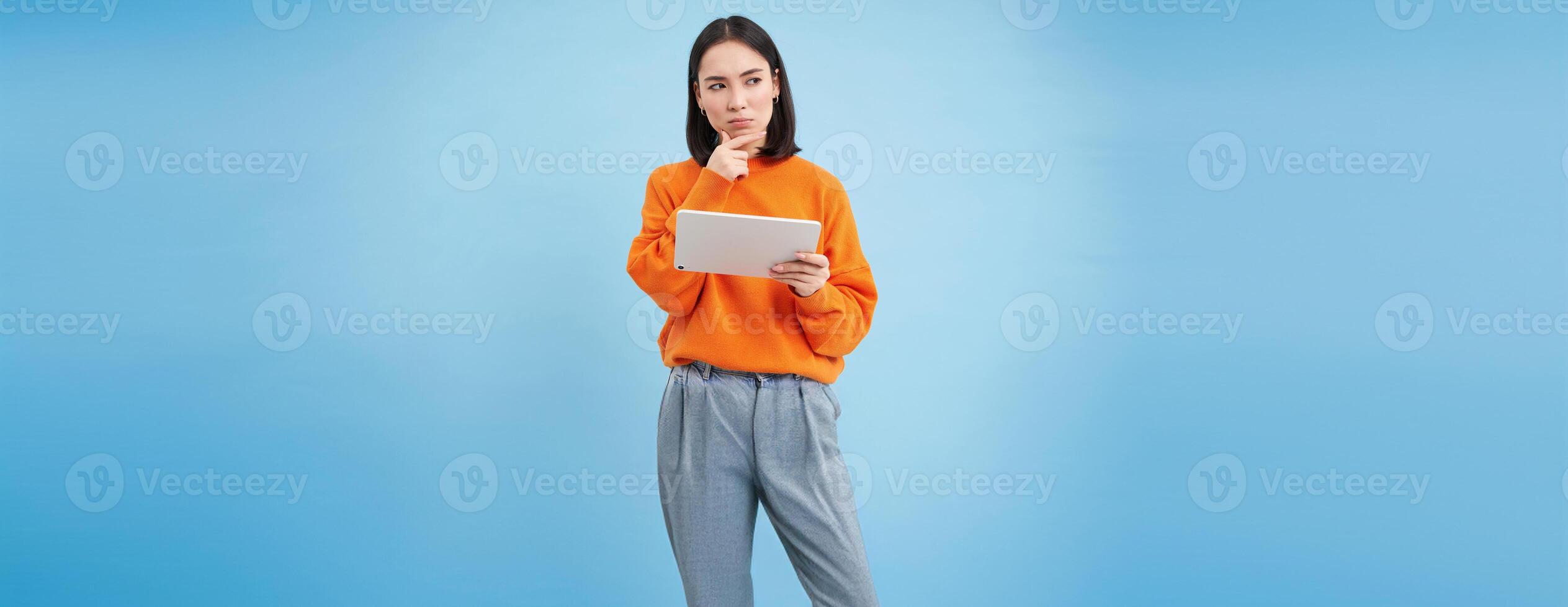 Woman with digital tablet thinking, looking aside with thoughtful face expression, standing over blue background photo