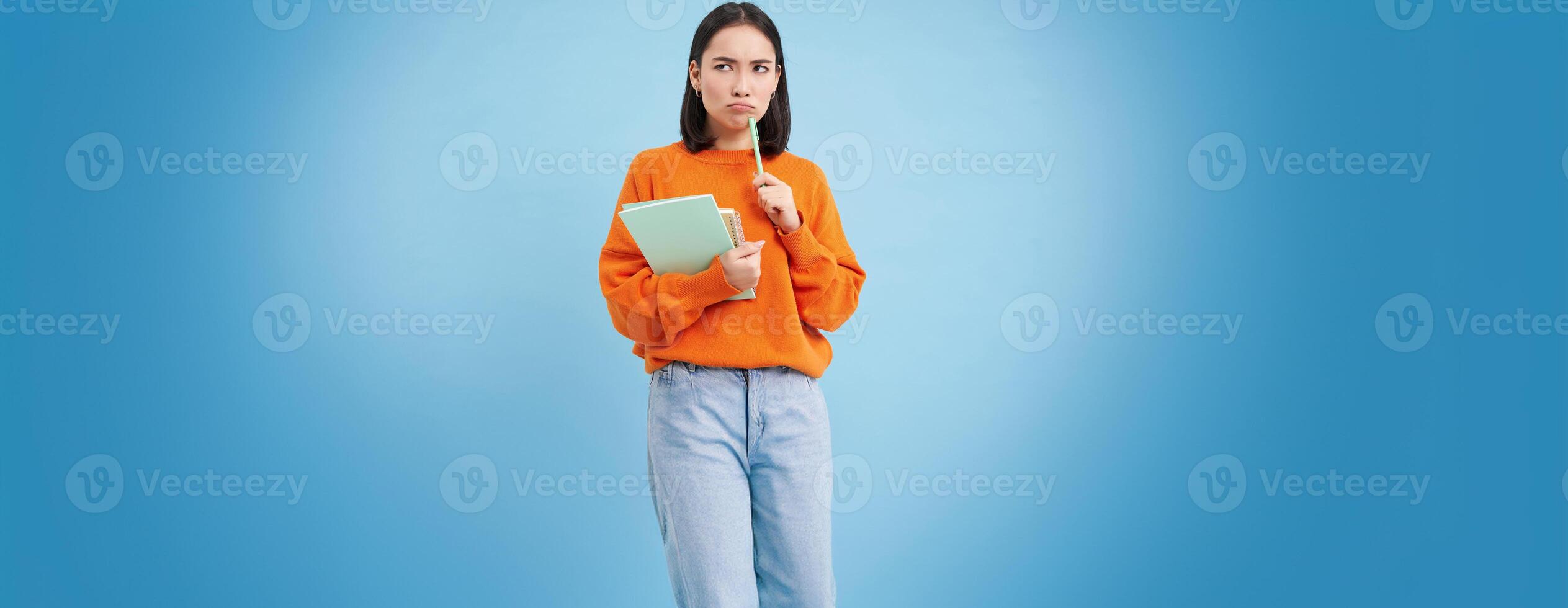 Asian girl, student with thinking face, pondering smth with serious face, holding pencil and notebooks, standing over blue background photo