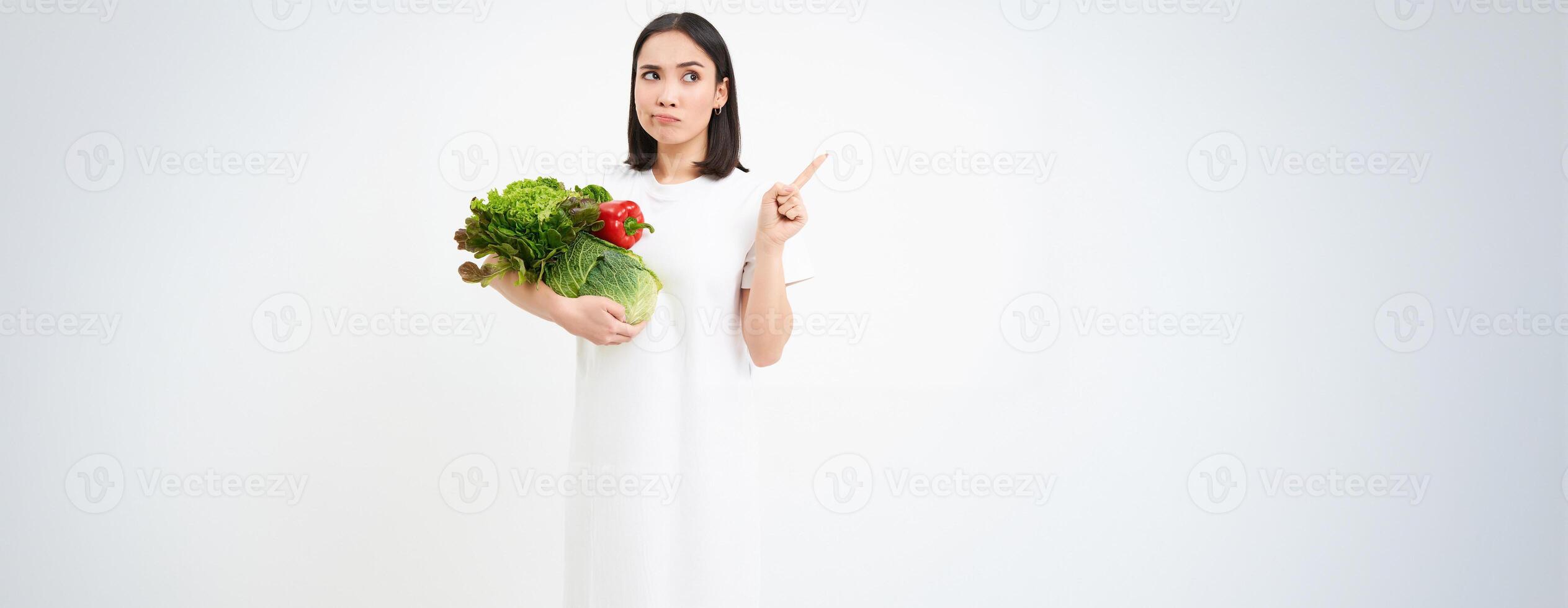 Portrait of pensive asian woman, holding vegetables, pointing right with thinking face, white background photo