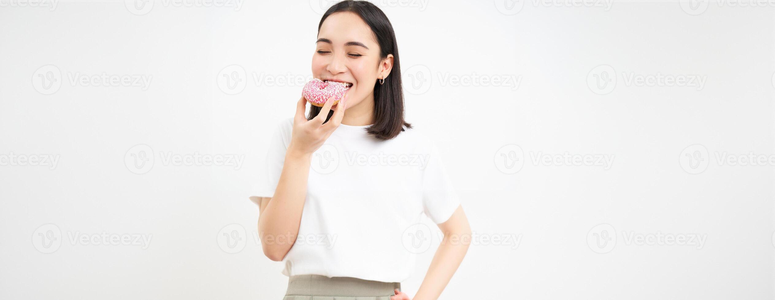 Close up of smiling pleased asian woman, eats glazed pink doughnut, enjoys eating tasty donnut, white studio background photo