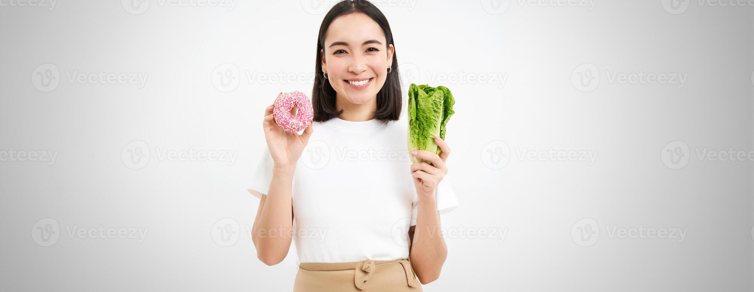 Healthy lifestyle concept. Happy smiling asian woman, showing one glazed doughnut and cabbage, giving choice, white background photo