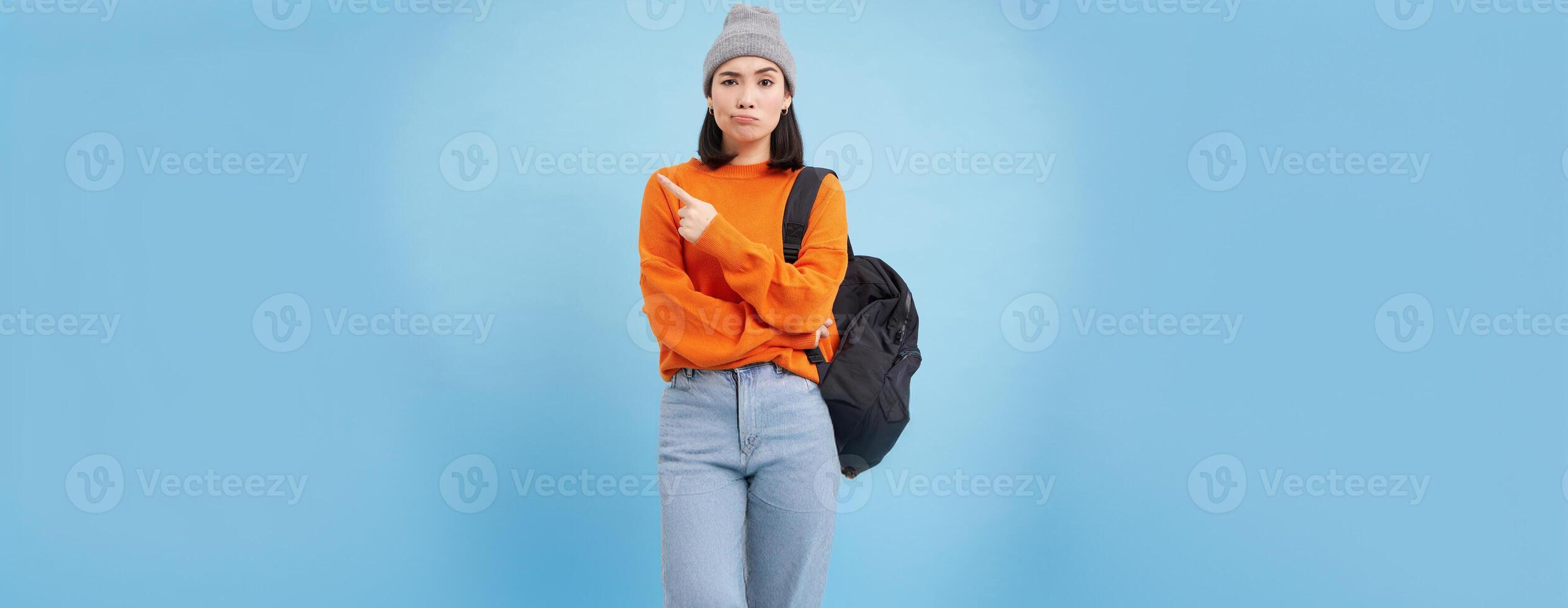 Skeptical asian woman pointing left at strange banner, looks doubtful, stands over studio background photo