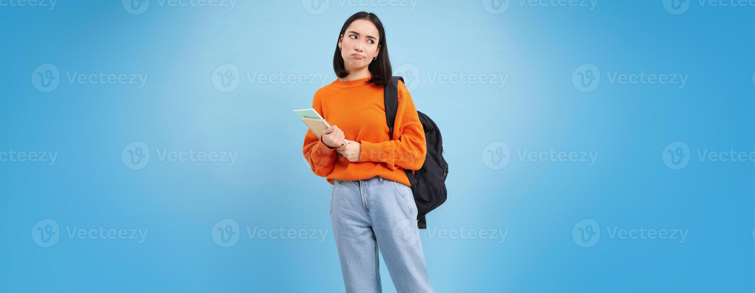 Bored college student, girl with backpack and notebooks, sulks and rolls eyes, stands upset against blue background photo
