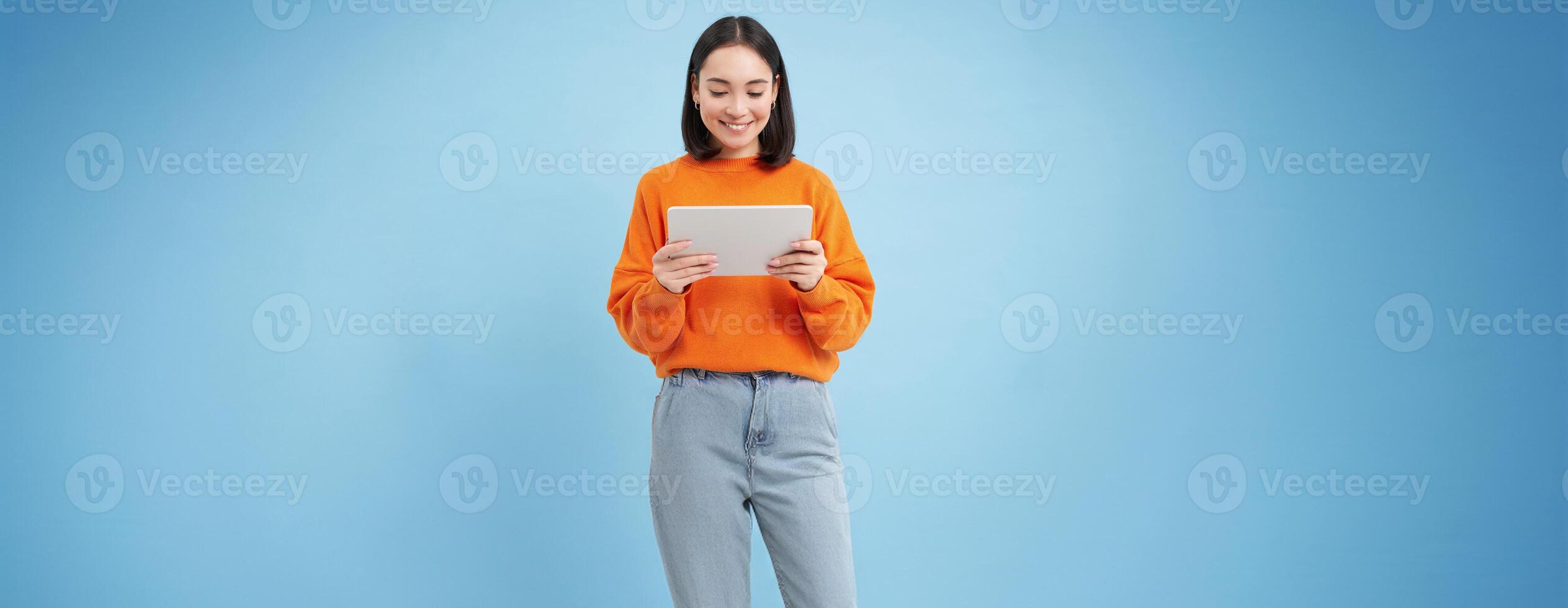 Smiling beautiful asian woman reading on digital tablet, watching videos on gadget or playing games, standing over blue background photo
