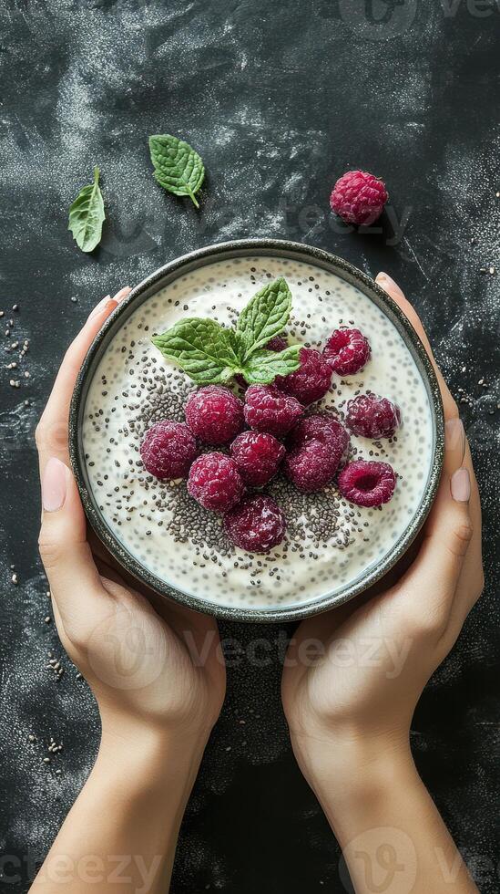 Delicious bowl of yogurt topped with fresh raspberries and chia seeds presented by hands on a textured black background photo