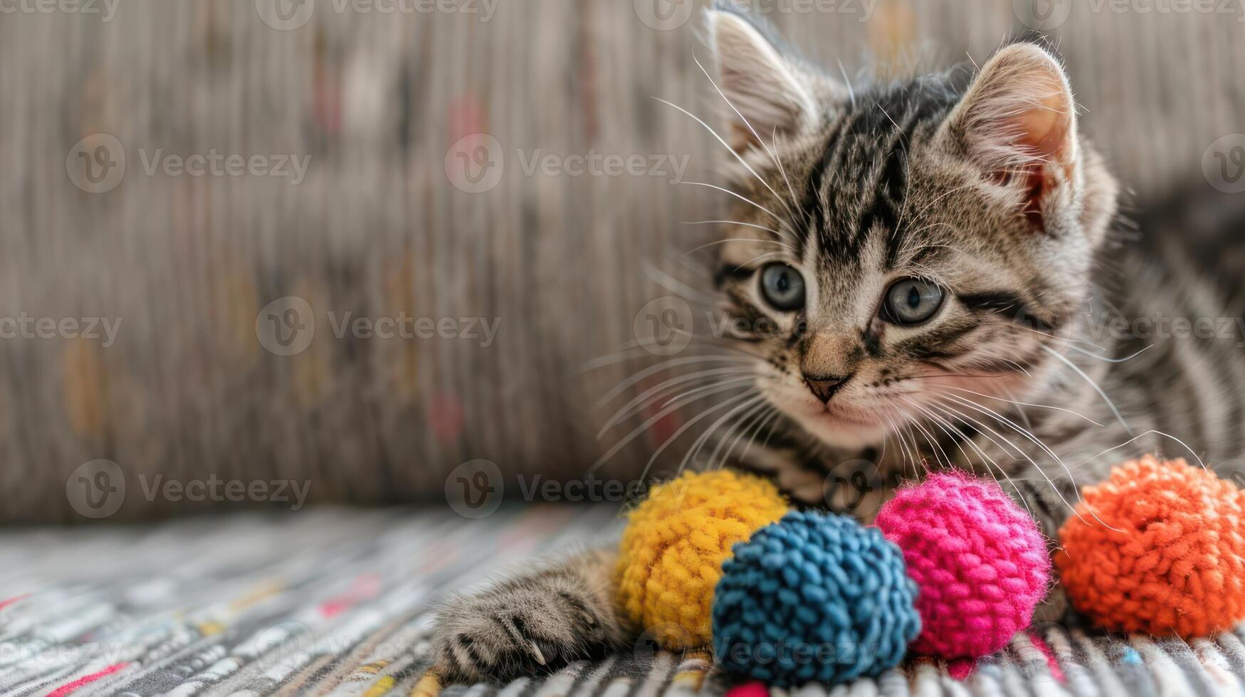 A playful kitten rests beside colorful yarn balls on a patterned surface. photo