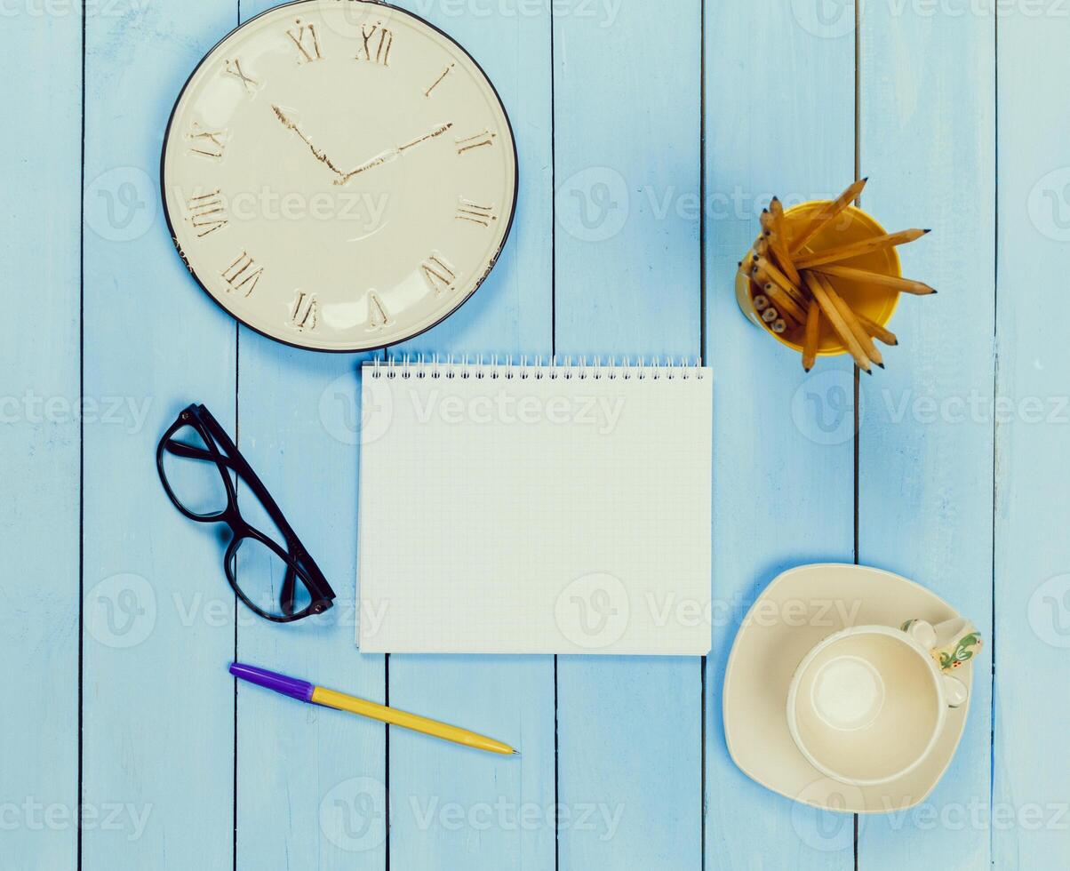 Workspace setup with a clock, notepad, glasses, a pen, a plant, and a coffee cup on a blue wooden table photo